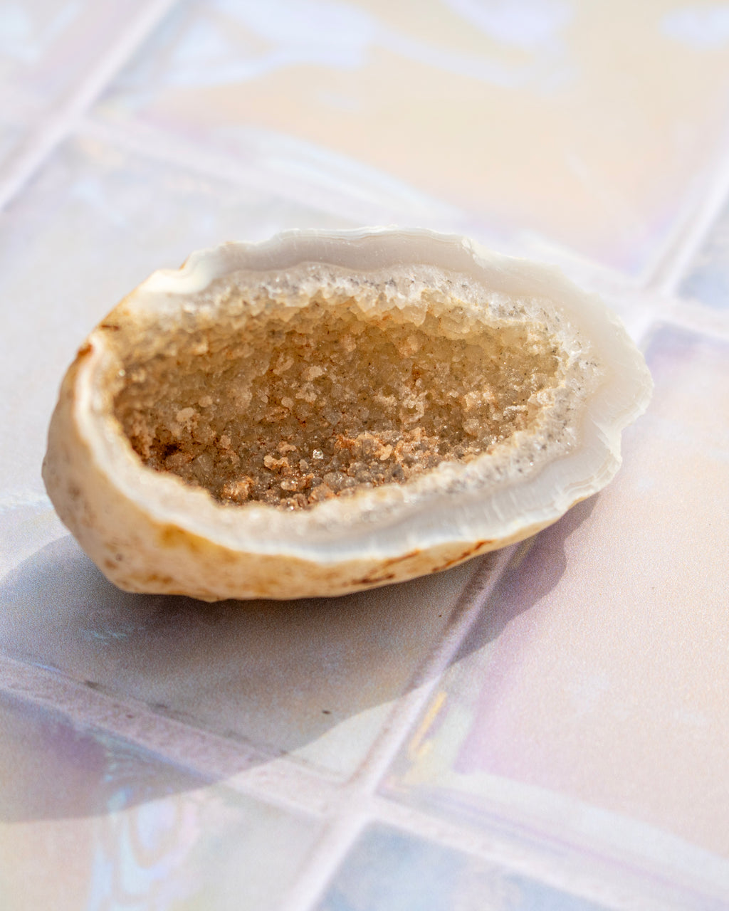 A geode shell half-shell filled with natural sand or sugar crystals, resting on a glossy, iridescent tiled surface.