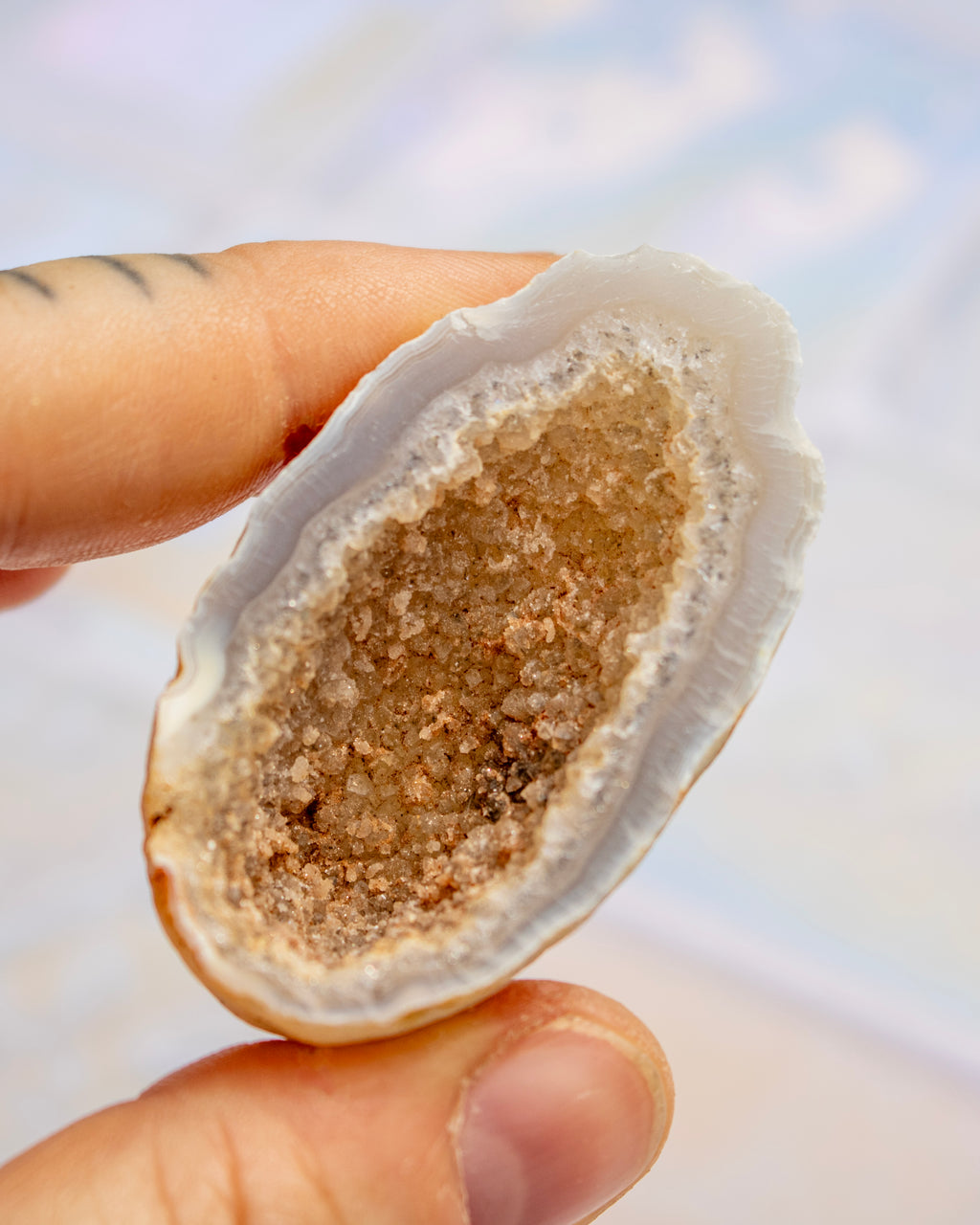 A close-up of a hand holding a geode specimen, showing a textured, crystalline beige and brown interior against a soft, blurred beach background.