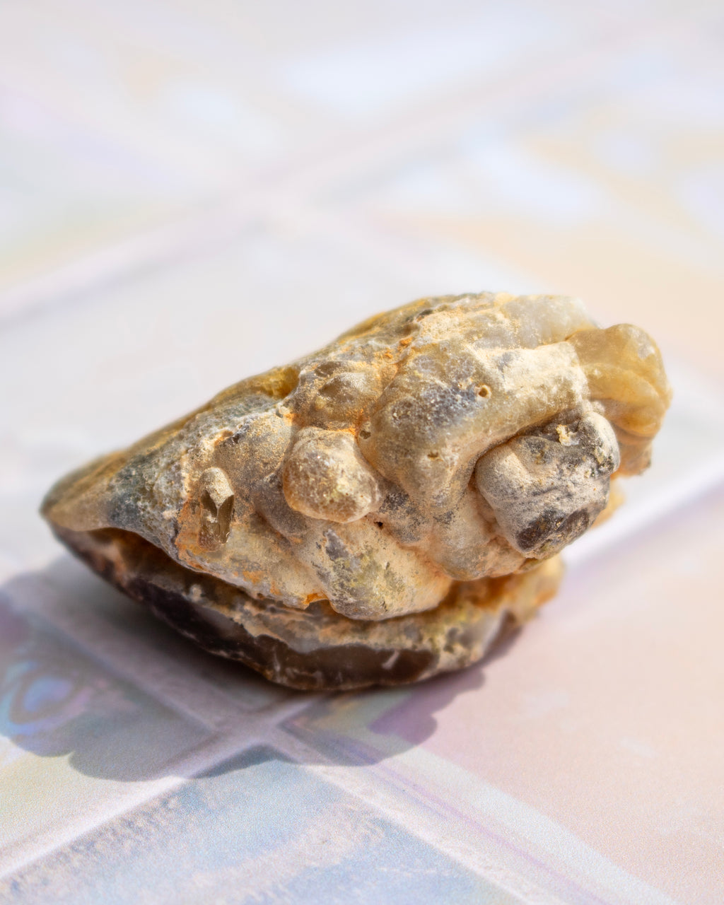 A small natural stone or mineral specimen with a rough, textured surface in beige, brown, and gray tones, photographed close-up on a softly blurred pastel surface.