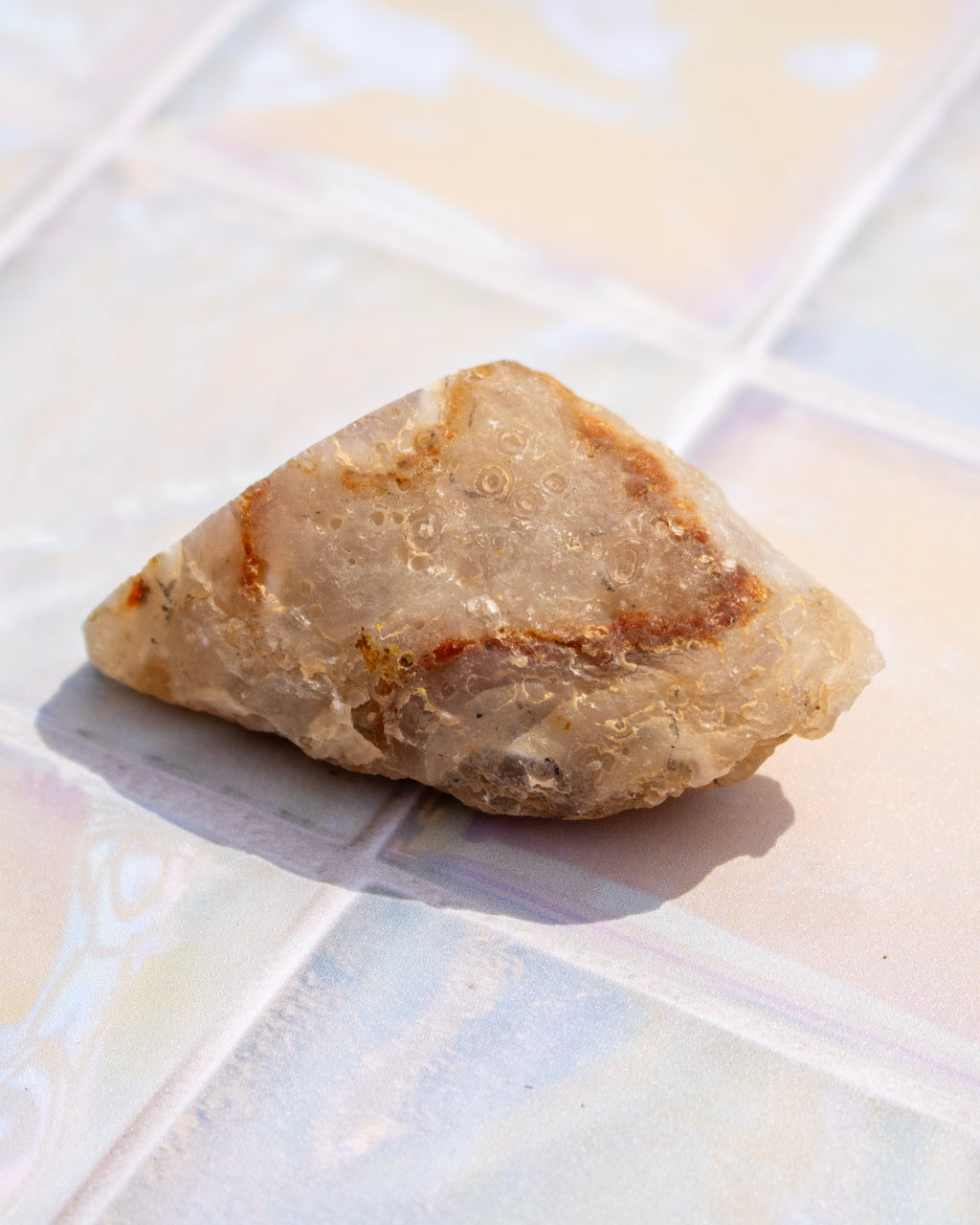 A rough, triangular piece of natural quartz or crystal with beige and light brown tones, resting on a glossy, iridescent tiled surface.