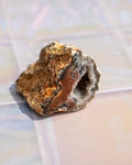 A natural mineral specimen or geode-like rock chunk with a rough crystalline texture, showing brown, orange, gray, and white tones, photographed on a pale tiled surface.