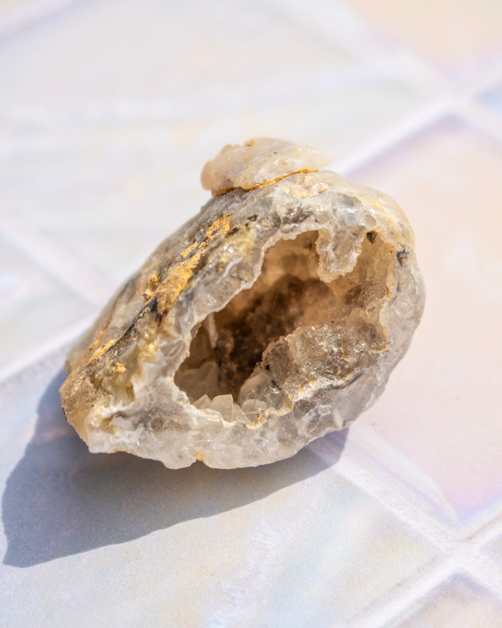 A raw crystal geode specimen with a hollow interior, featuring natural quartz-like formations in off-white, beige, and light brown tones, resting on a glossy white tiled surface.