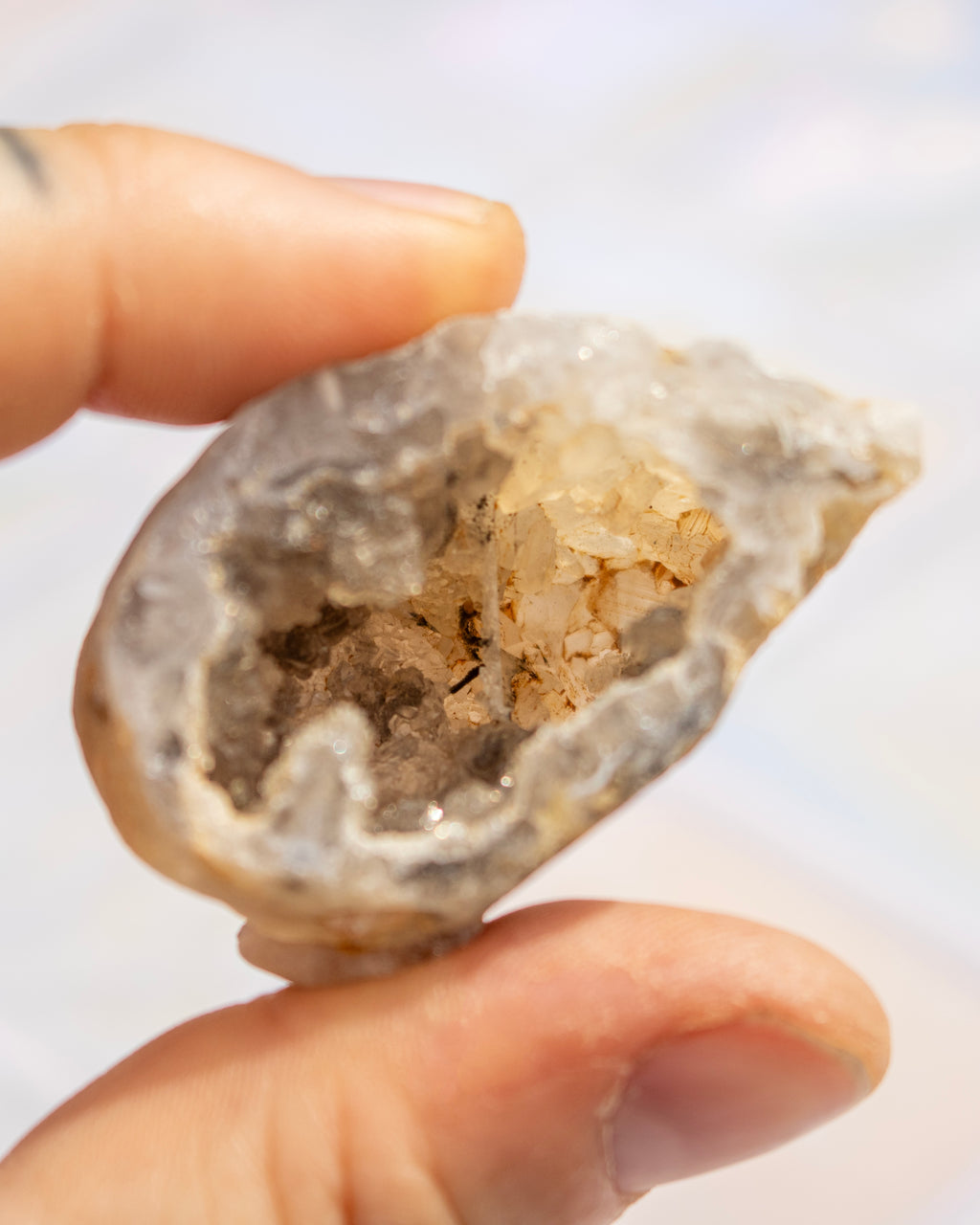 A close-up of a hand holding a raw crystal geode specimen with a hollow interior filled with pale quartz crystals against a soft, out-of-focus light background.
