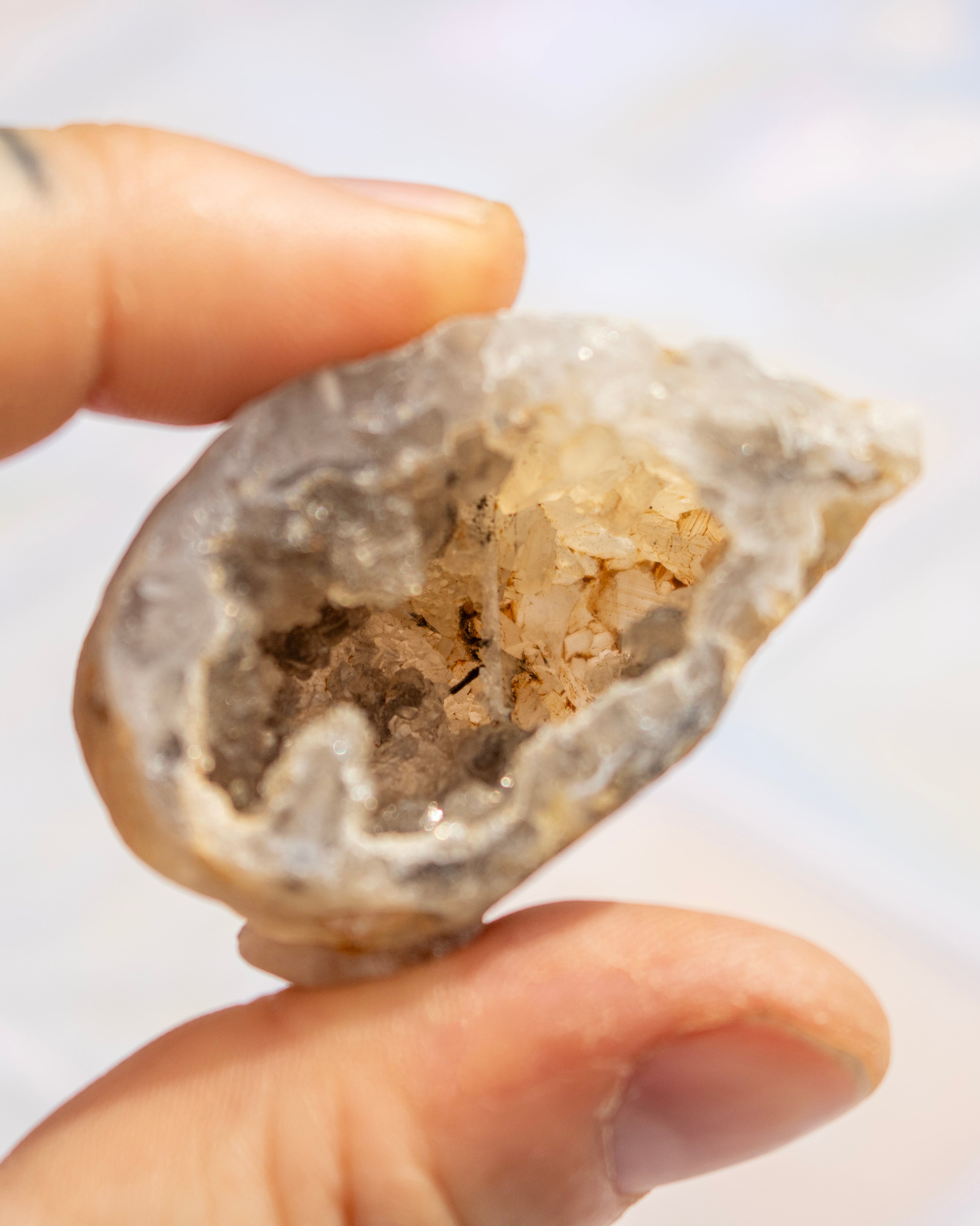 A close-up of a hand holding a raw crystal geode specimen with a hollow interior filled with pale quartz crystals against a soft, out-of-focus light background.