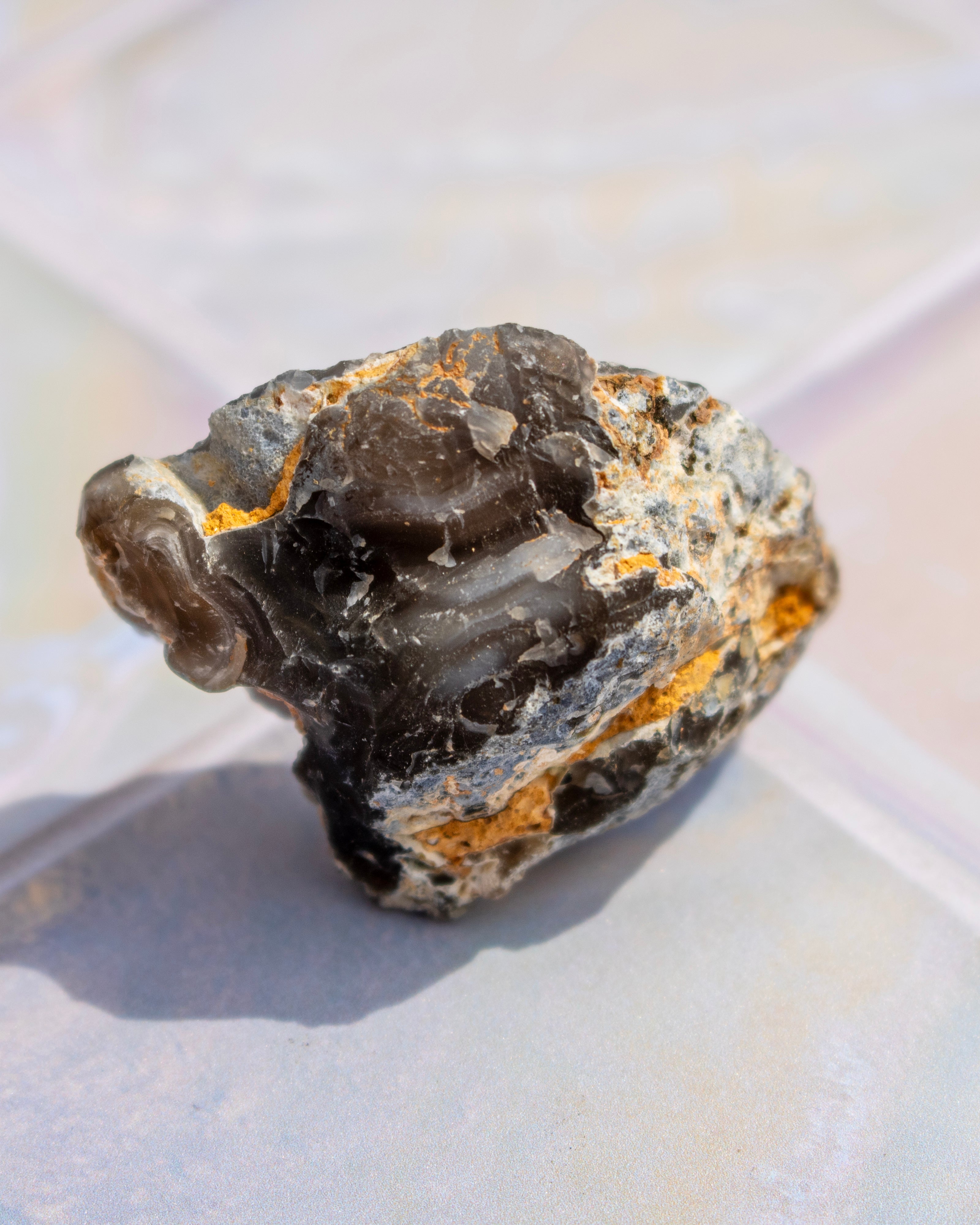 A rough, polished natural mineral specimen with dark gray to black smoky sections and bright orange-brown matrix, resting on a light gray surface.