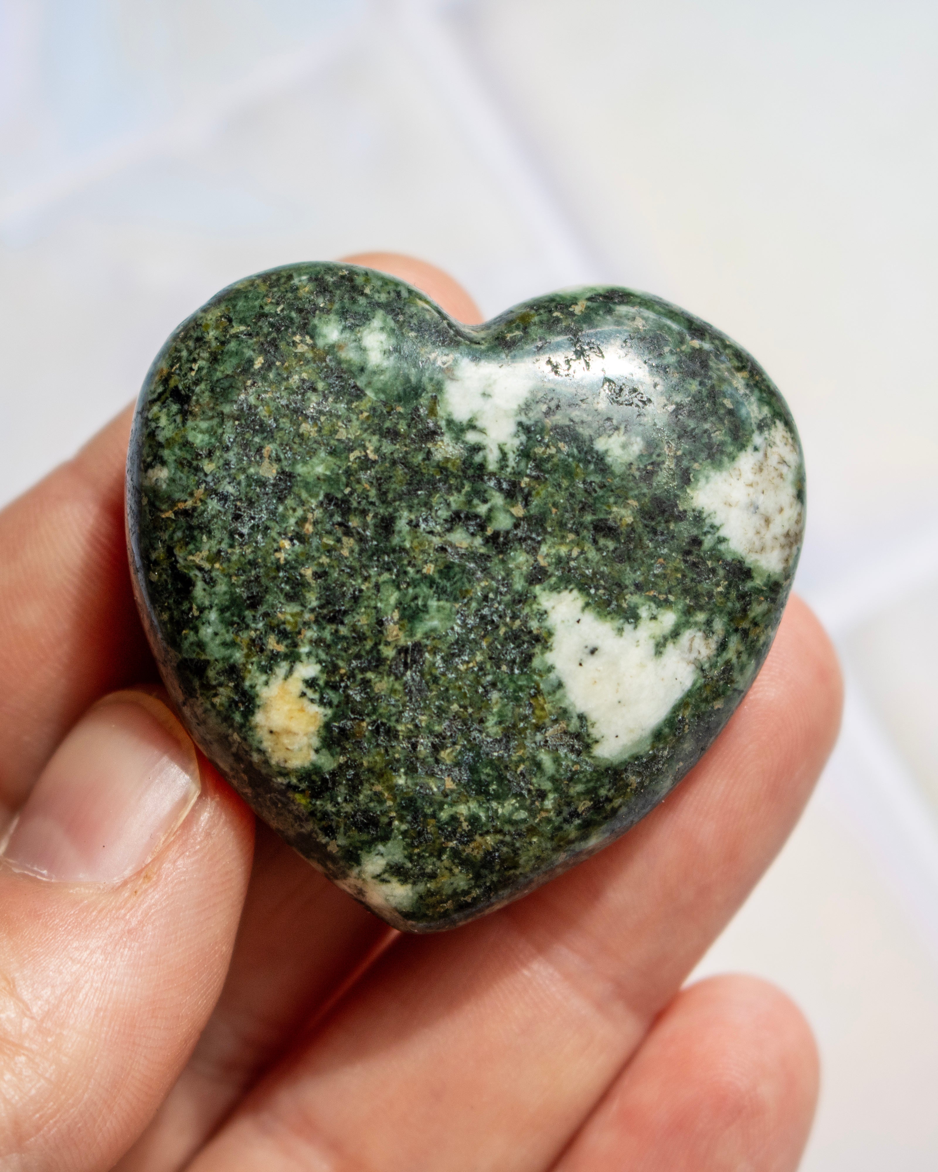 A heart-shaped polished stone with a mottled pattern of dark green, black, and white, held in a person's hand.