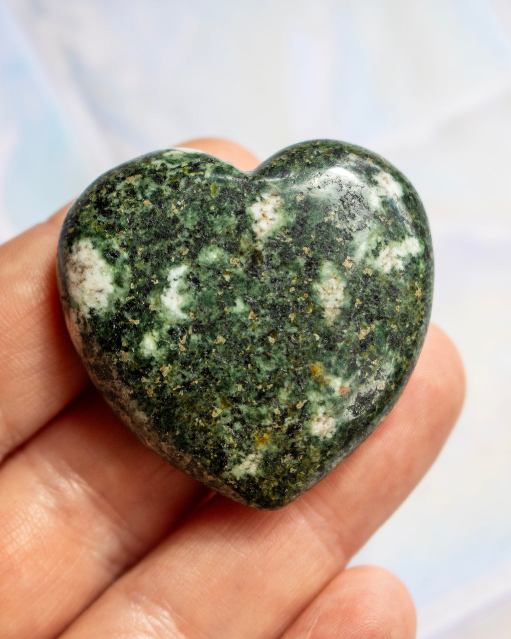 A polished heart-shaped green stone with white and dark speckled inclusions, held in a person's hand against a soft light blue background.