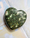 A polished heart-shaped green stone with natural white and dark mottled patterns, resting on a light-colored tiled surface.