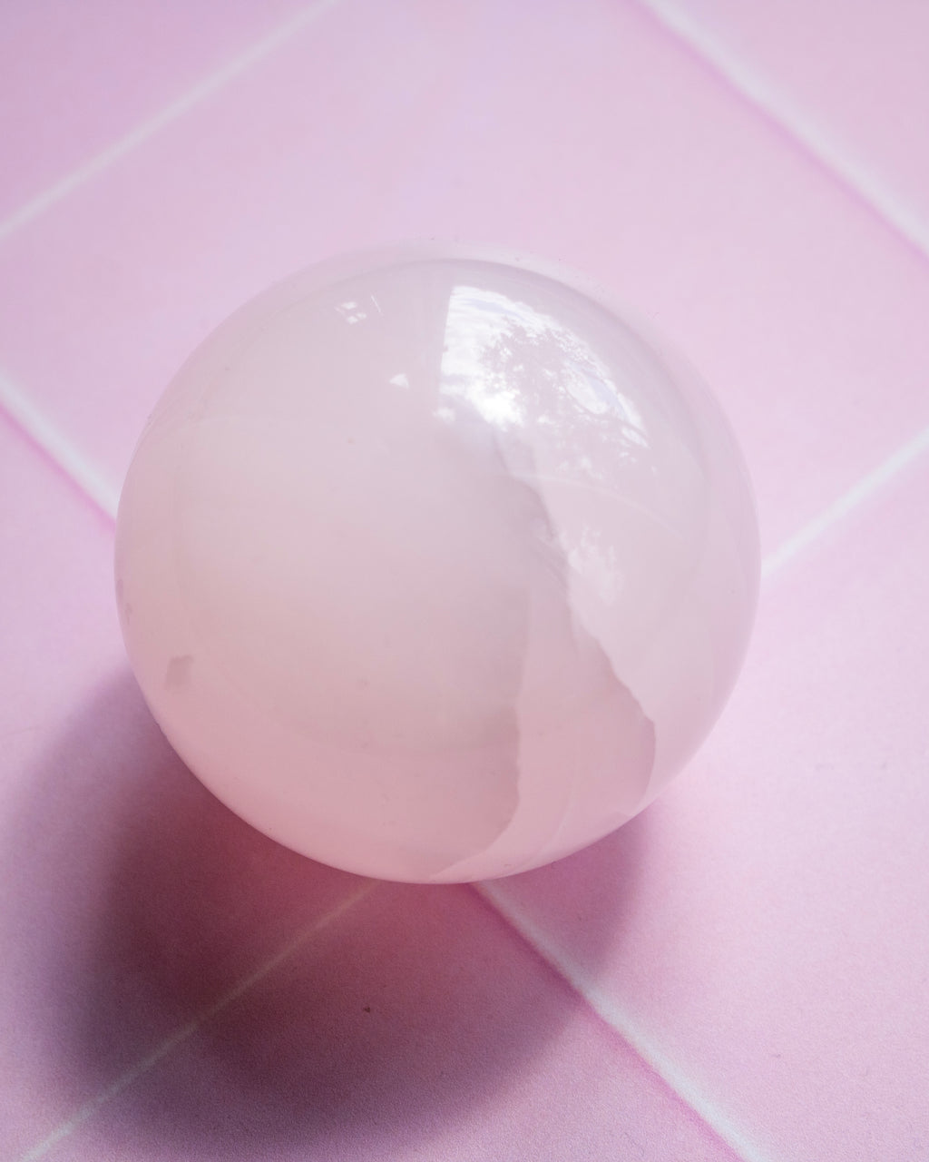 A smooth, polished pale pink crystal sphere resting on a pink surface with a soft shadow cast to the left.