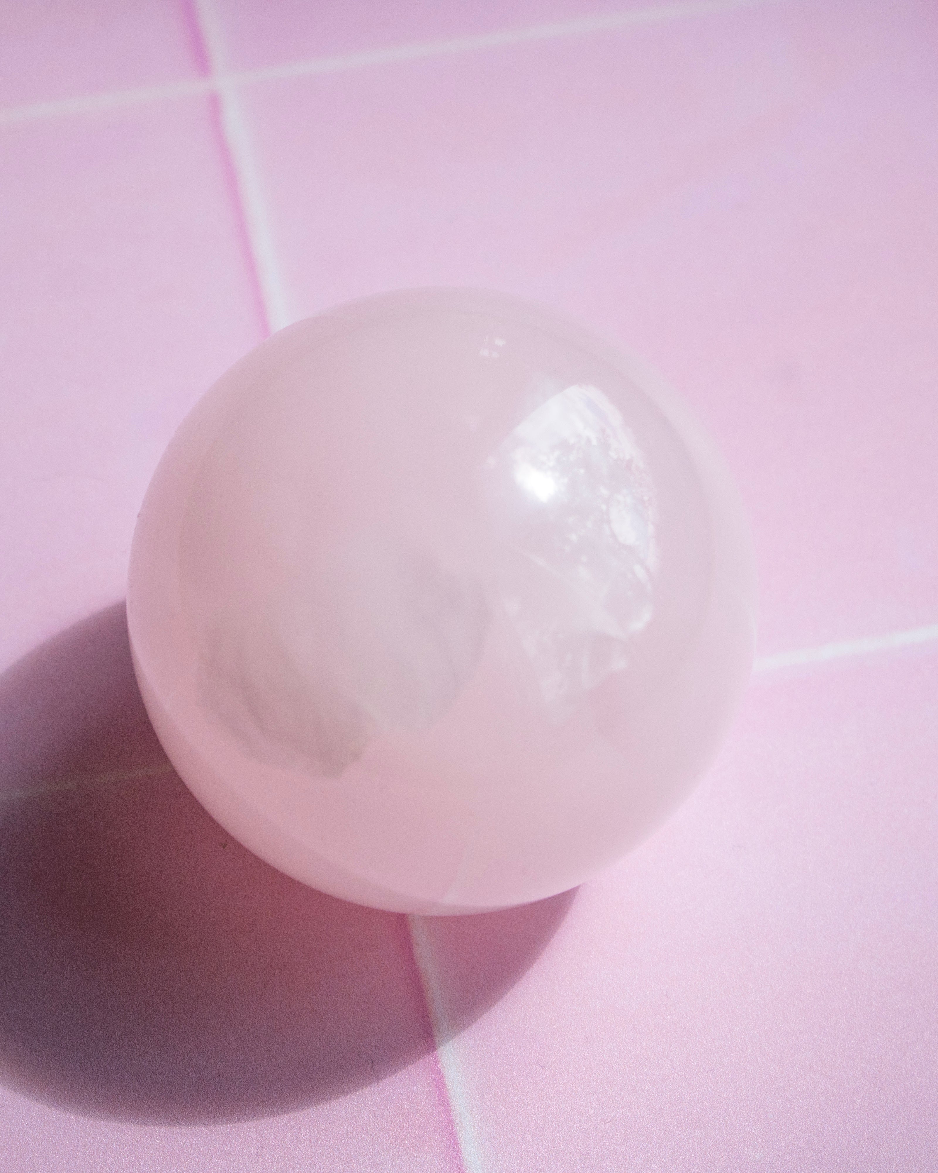 A smooth, polished pale pink sphere, resting on a pink tiled surface with a soft shadow cast to the left.