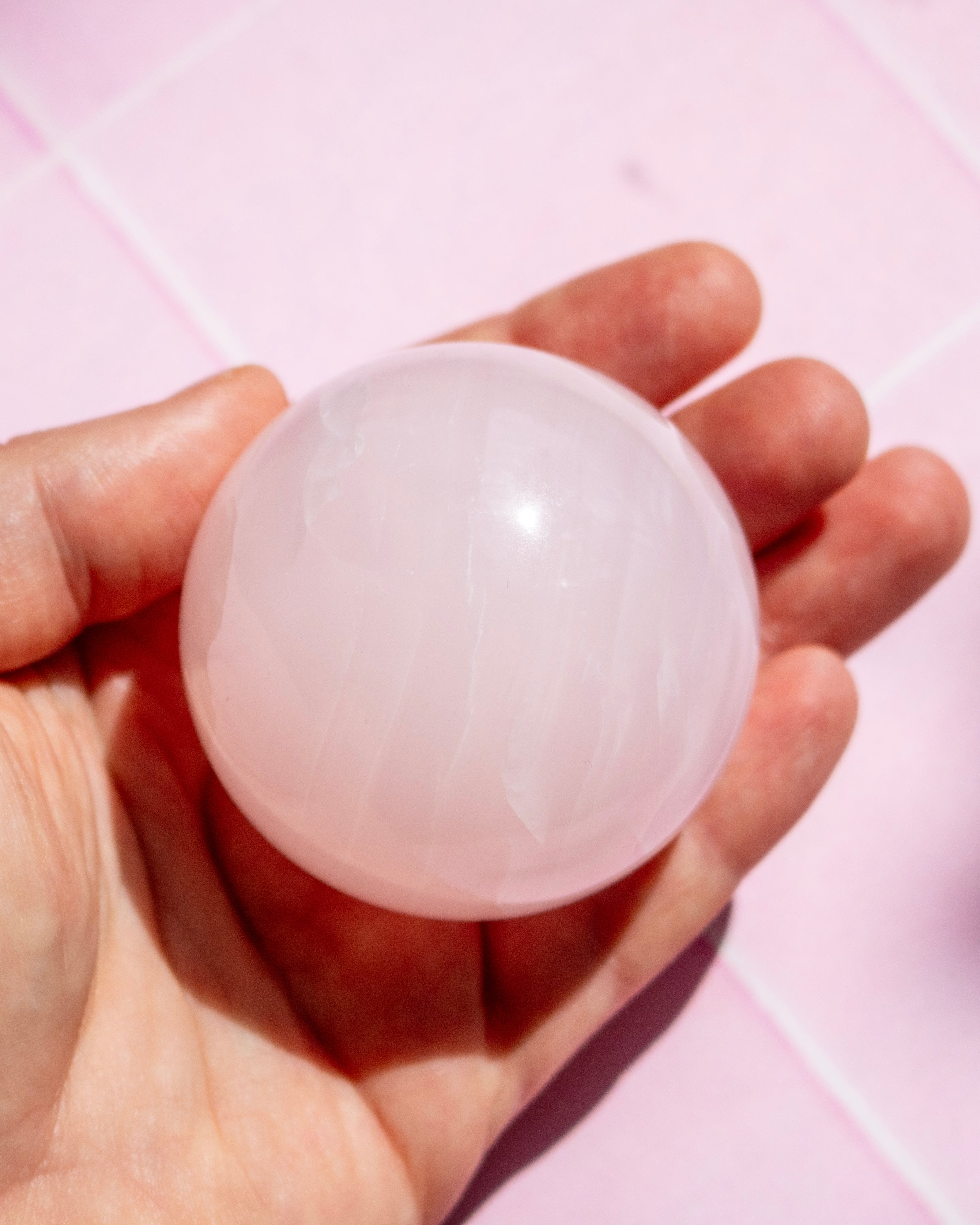 A hand holding a smooth, translucent pale pink sphere against a soft pink tiled background.