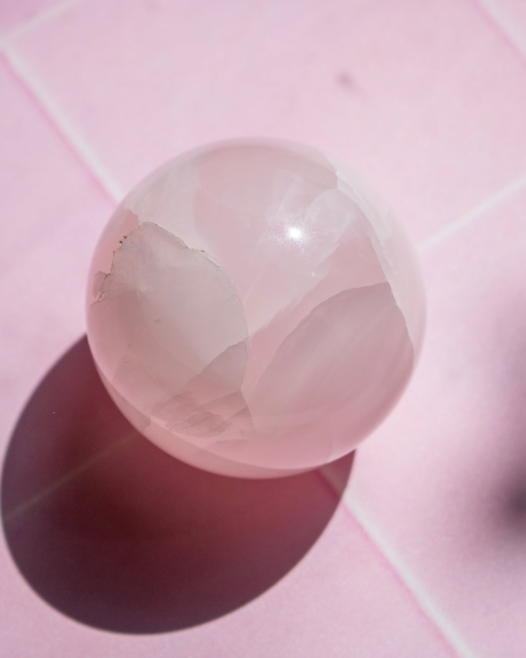A polished pink stone sphere resting on a pink tiled surface, casting a dark shadow to the left.