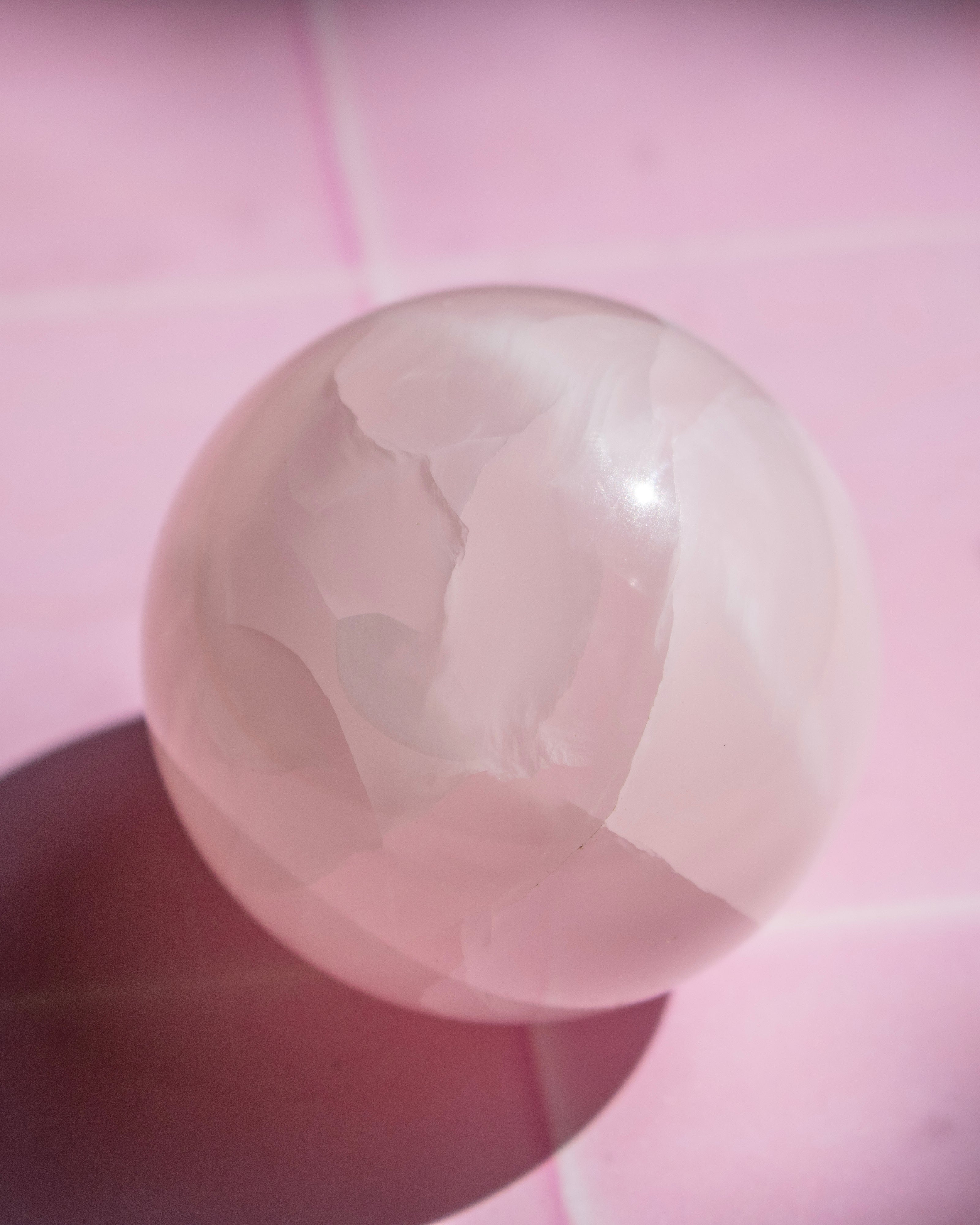 A polished pale pink stone sphere with natural marbled veining, resting on a pink surface with a soft shadow.