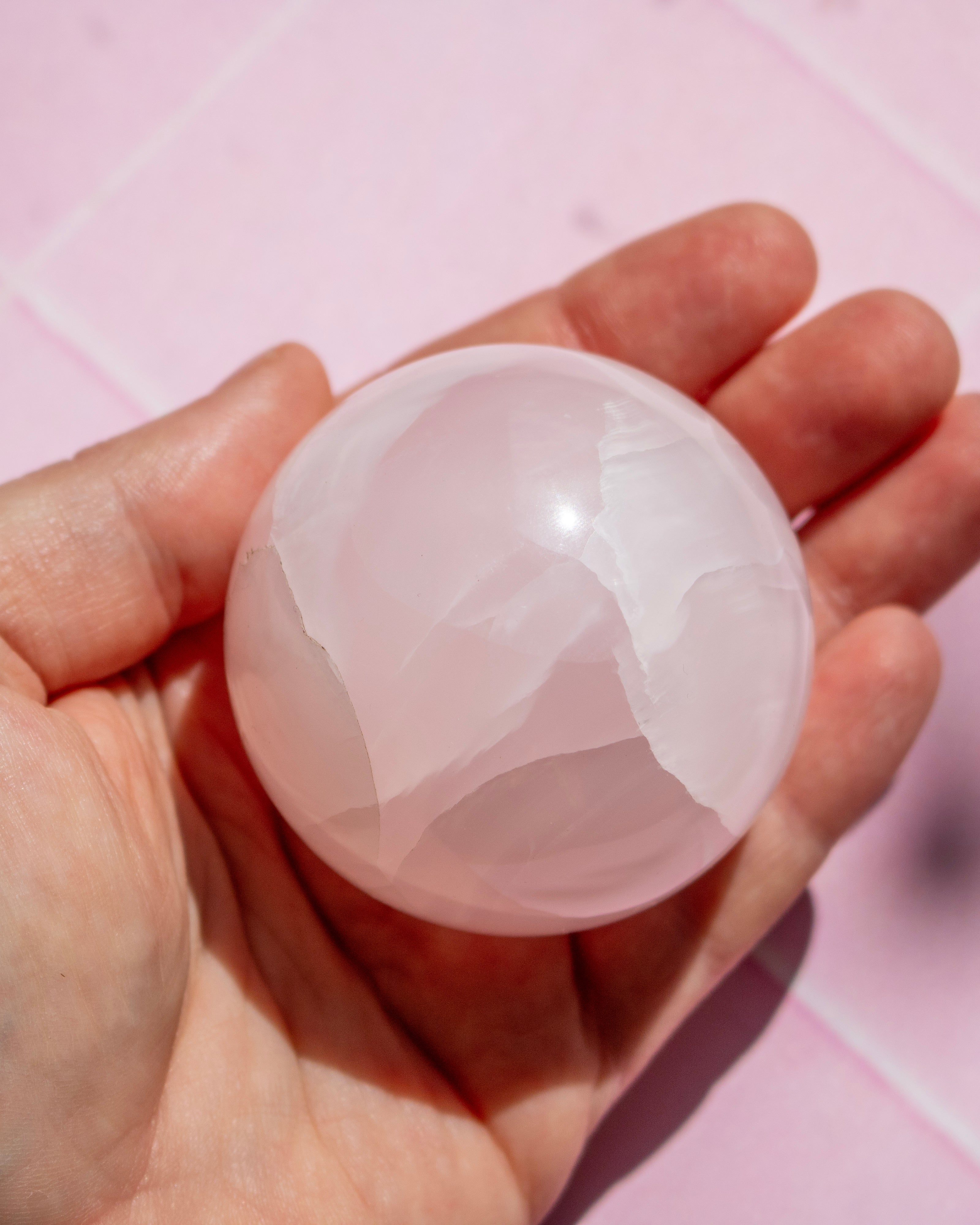 A hand holding a smooth, polished pink stone sphere against a soft pink background.