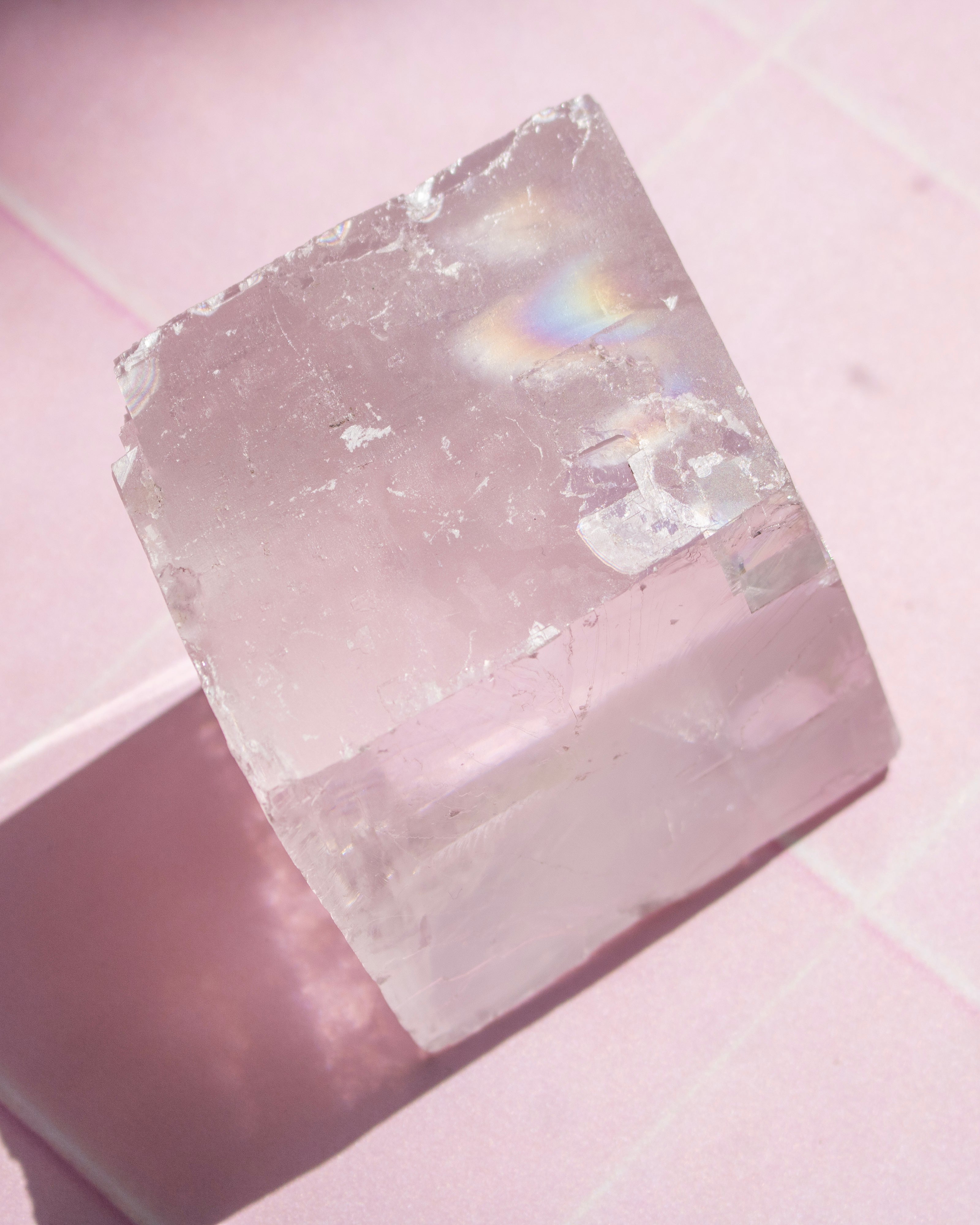 A translucent pale pink crystal or gemstone block photographed on a pink surface, with soft light and a faint iridescent rainbow reflection near the top.