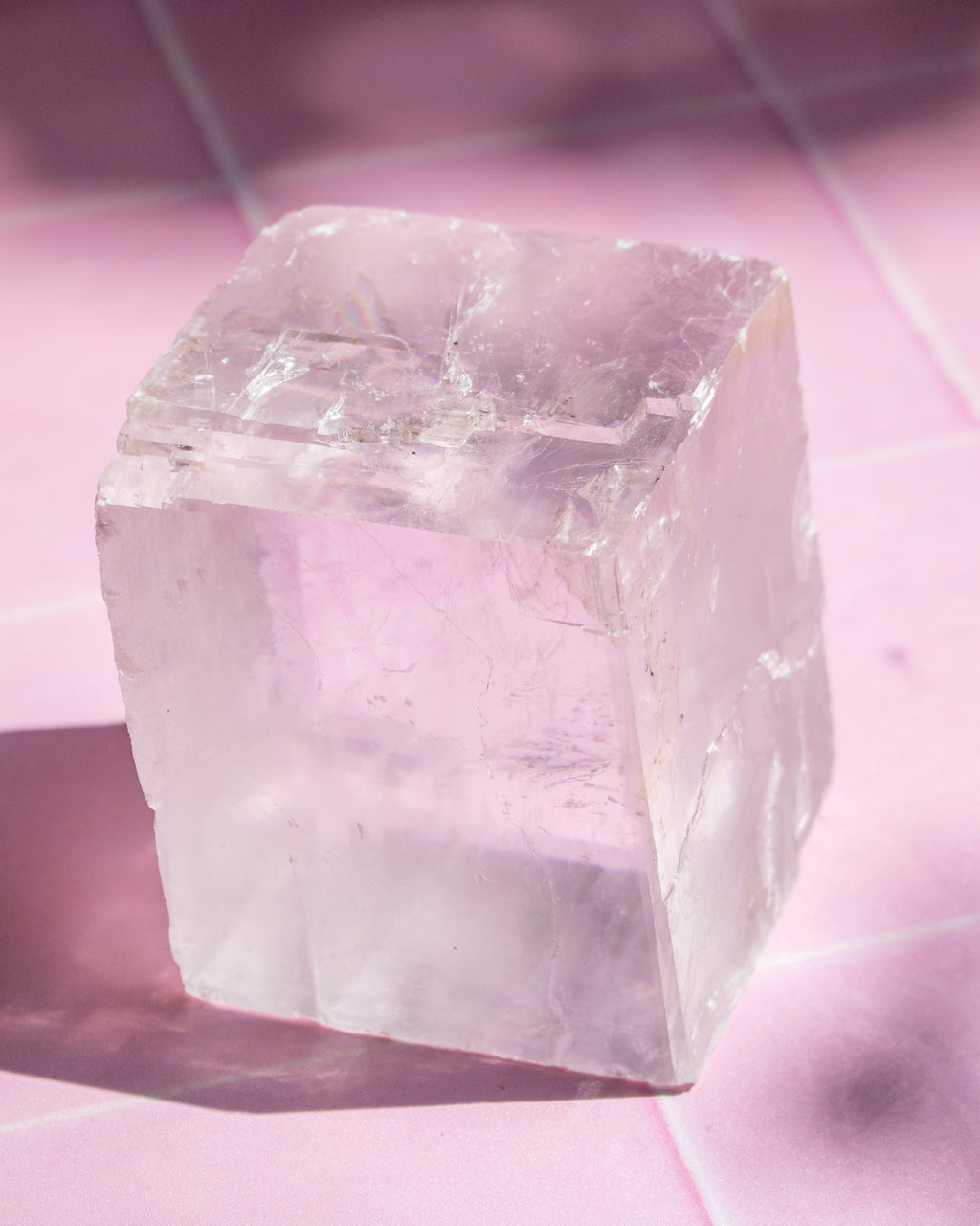 A translucent pale pink crystal cube with rough, faceted edges, photographed on a pink surface with strong sunlight and shadows.