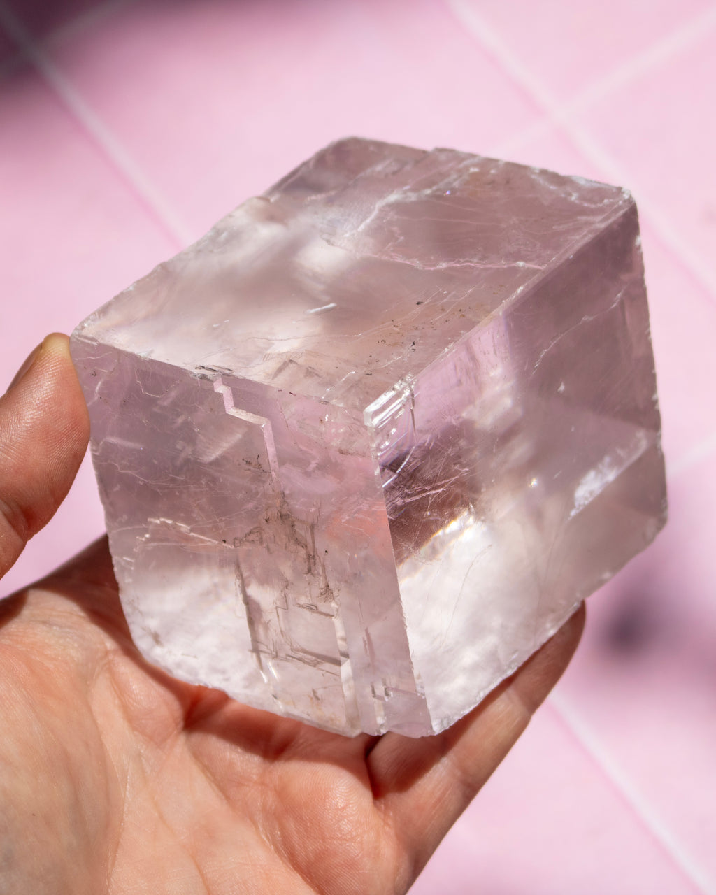 A hand holding a large, rough, translucent pale pink crystal cube against a soft pink background.