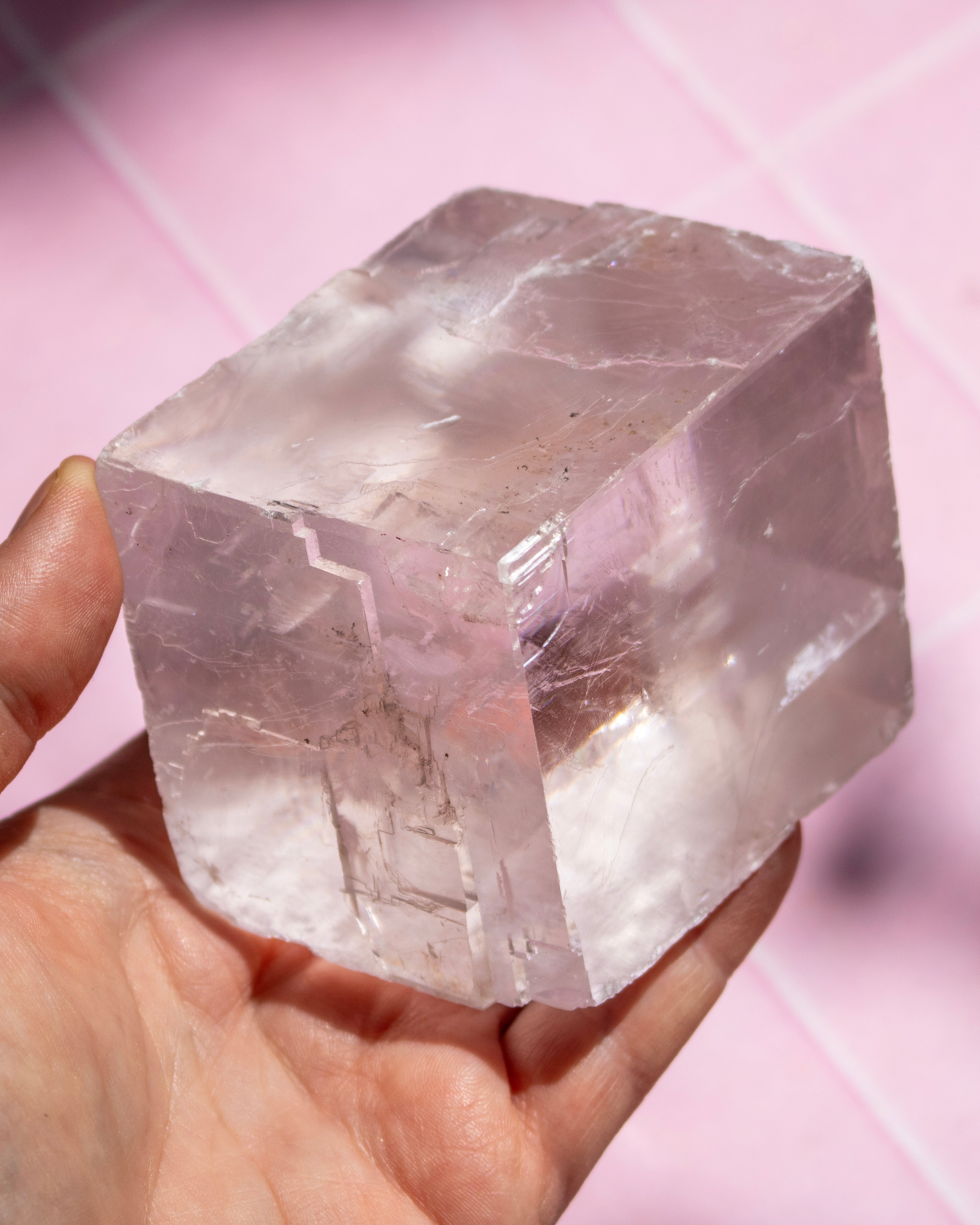 A hand holding a large, rough, translucent pale pink crystal cube against a soft pink background.