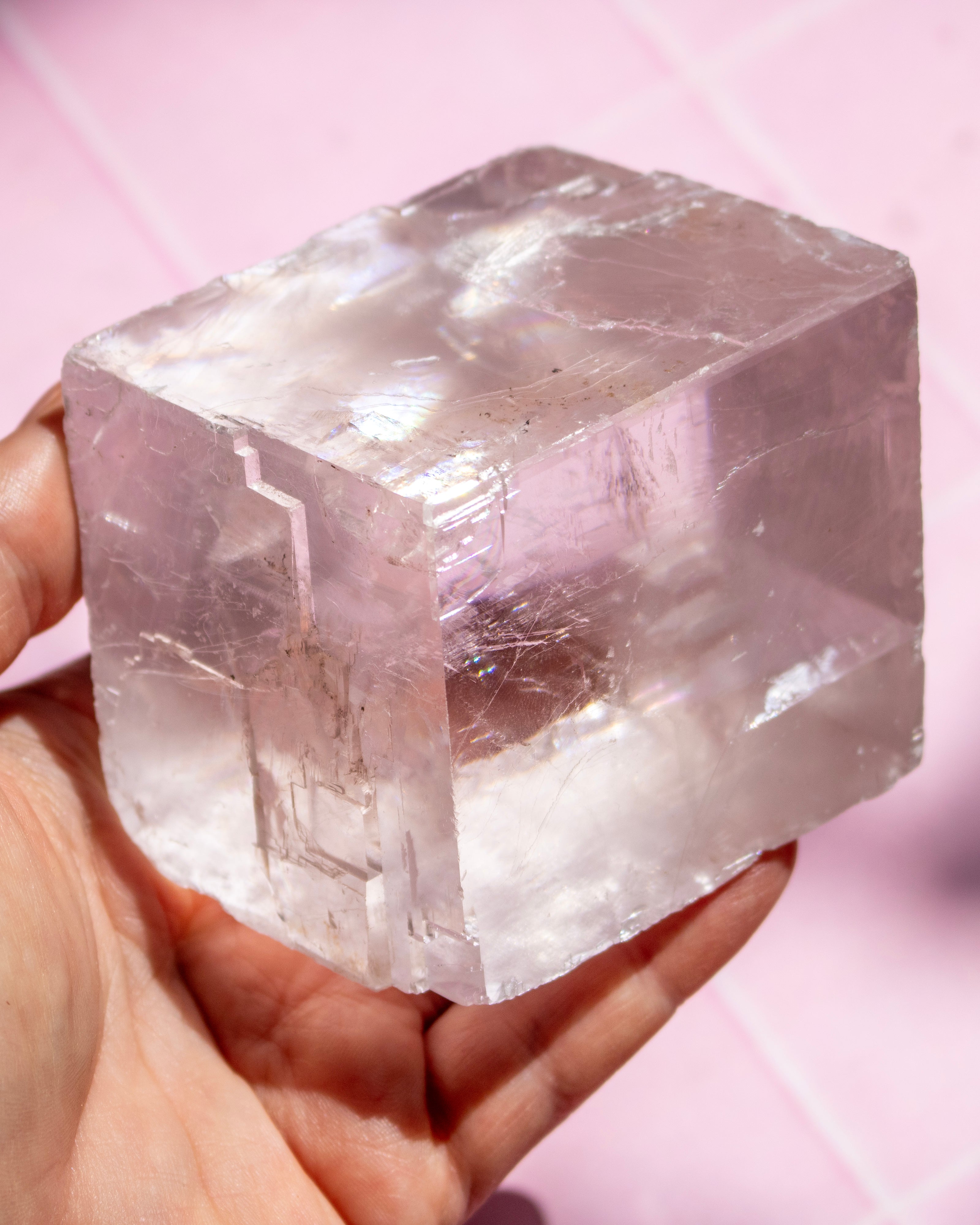 A hand holding a large, translucent pinkish-white crystal cube against a soft pink background.