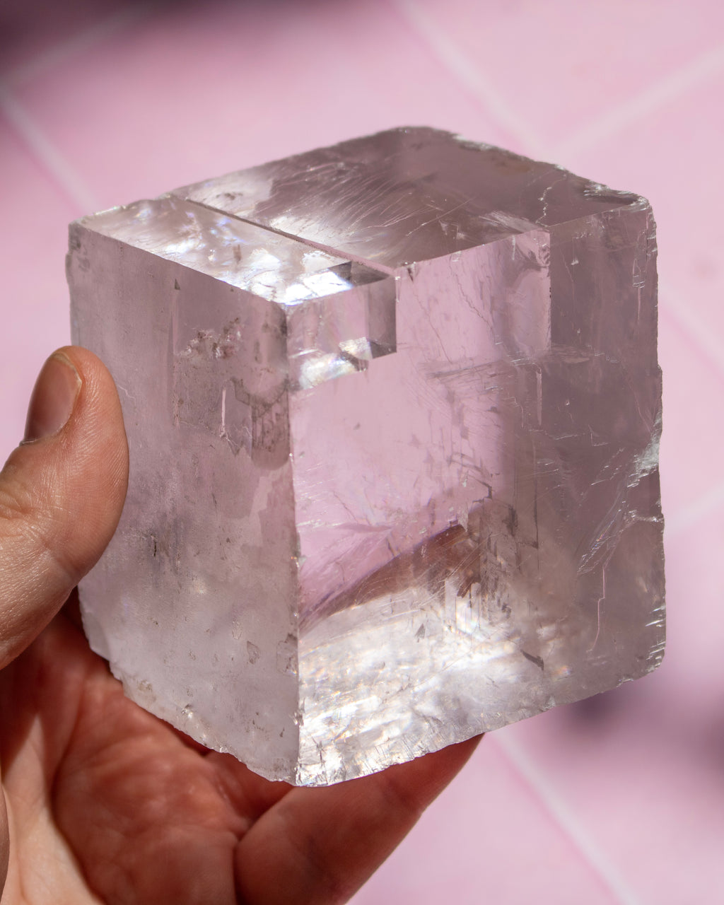 A hand holding a large, translucent pale pink crystal cube with visible internal inclusions and faceted edges against a soft pink background.