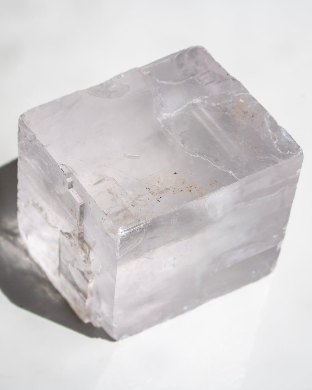A translucent clear crystal cube with natural rough edges and subtle internal inclusions, photographed on a white reflective surface with a soft shadow.