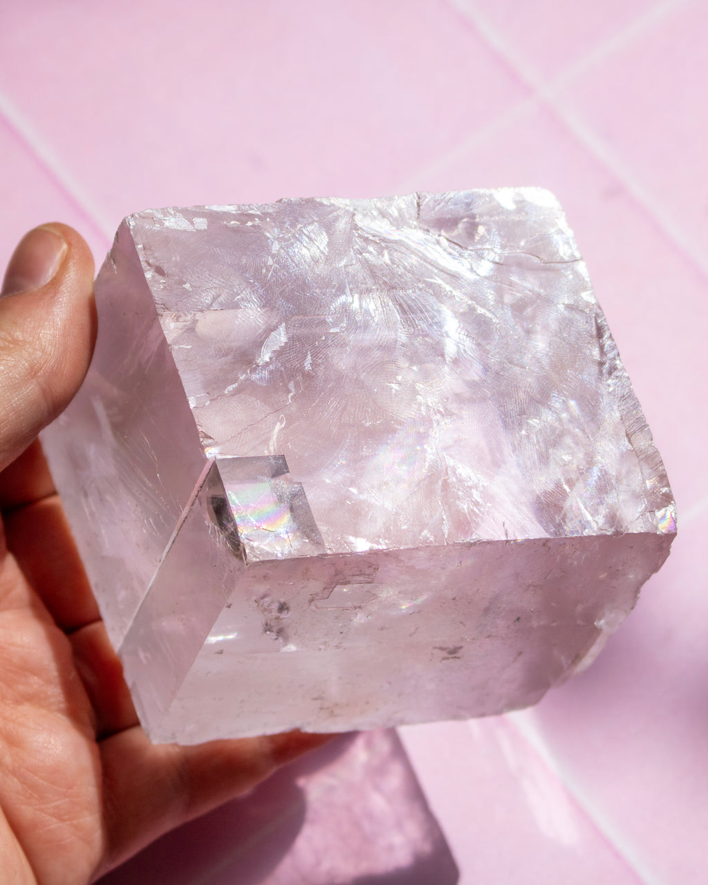 A hand holding a large, rough, translucent pink quartz crystal cube against a pink tiled background.