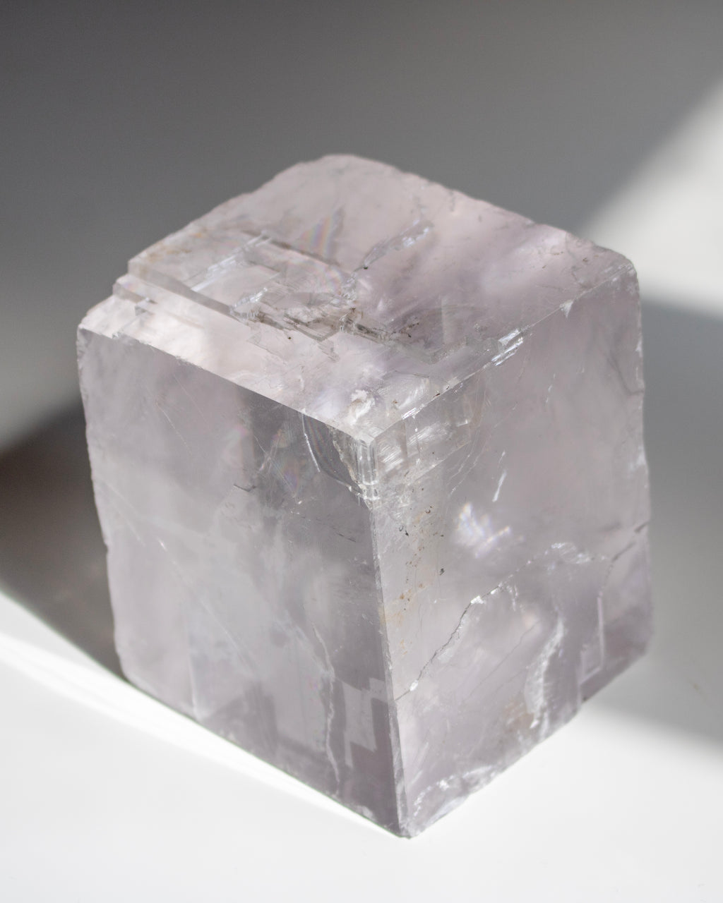 A translucent pale lavender crystal cube with natural rough edges and internal inclusions, photographed on a white surface with soft shadows.