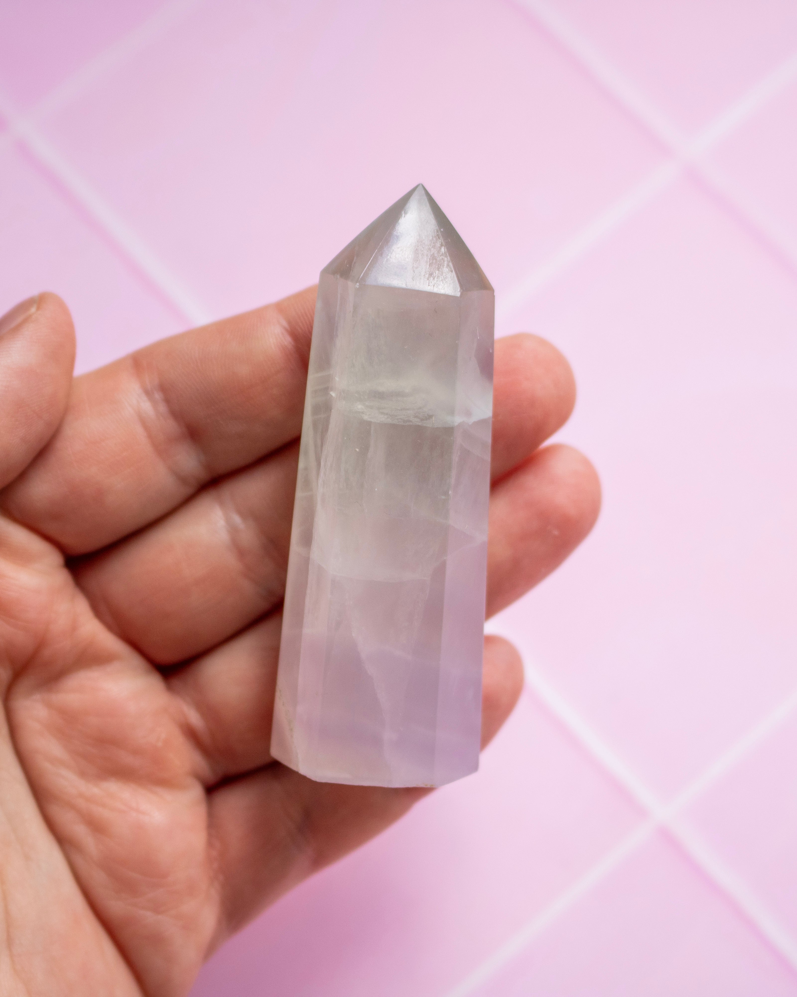 A hand holding a crystal point against a soft pink background with a faint grid pattern.
