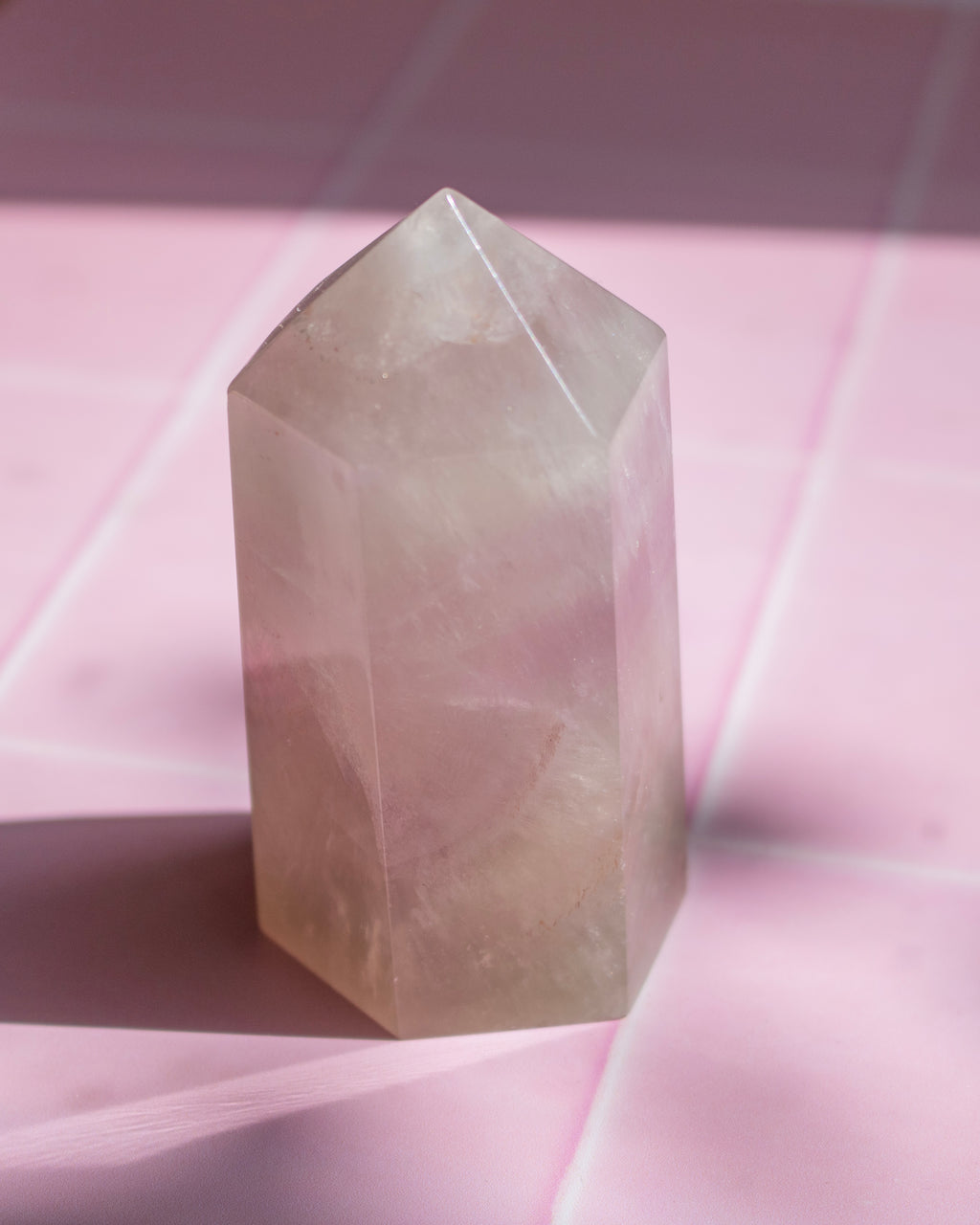 A pale pink translucent crystal tower with a pointed top, standing on a pink surface with soft shadows and a faint grid pattern in the background.
