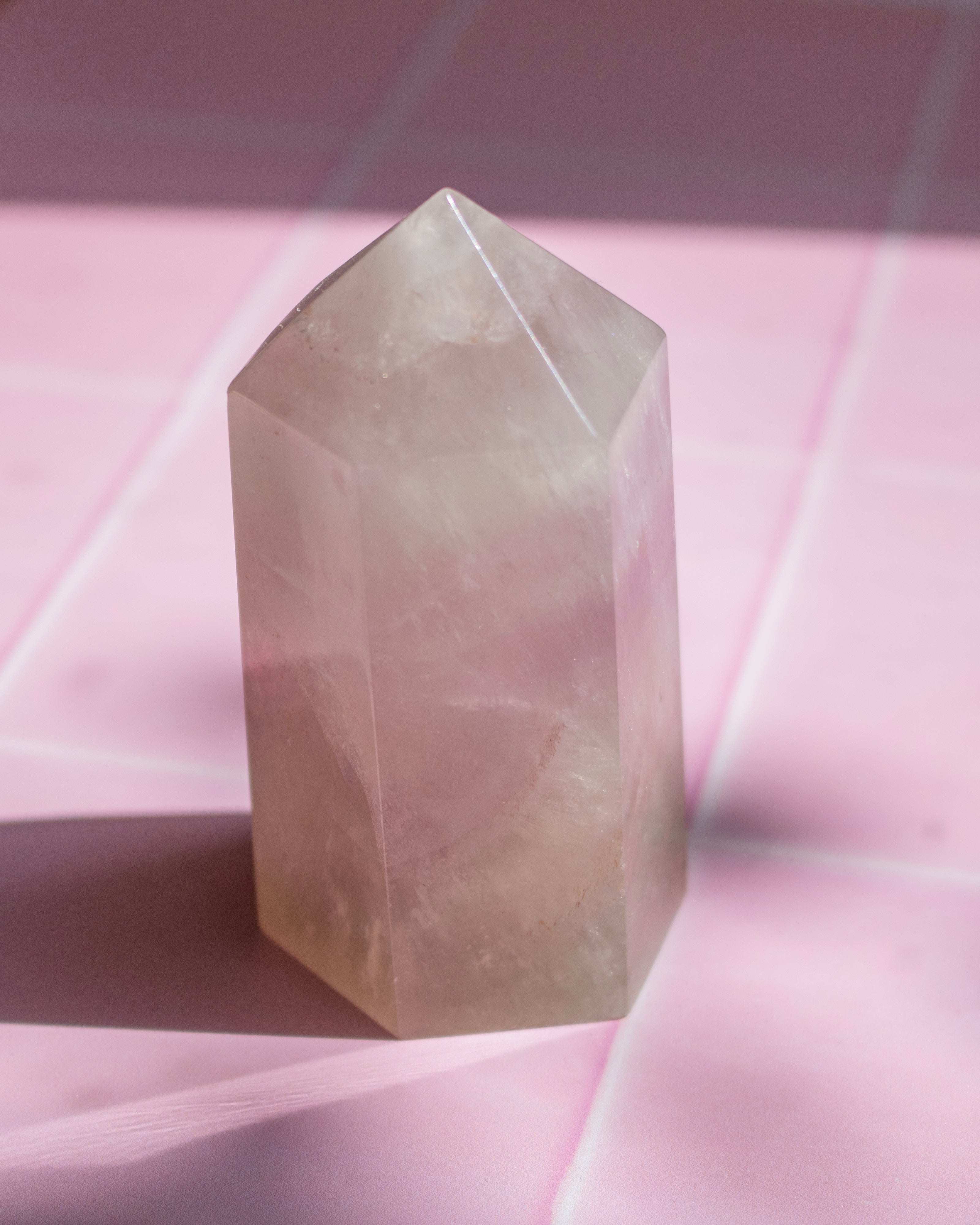 A pale pink translucent crystal tower with a pointed top, standing on a pink surface with soft shadows and a faint grid pattern in the background.