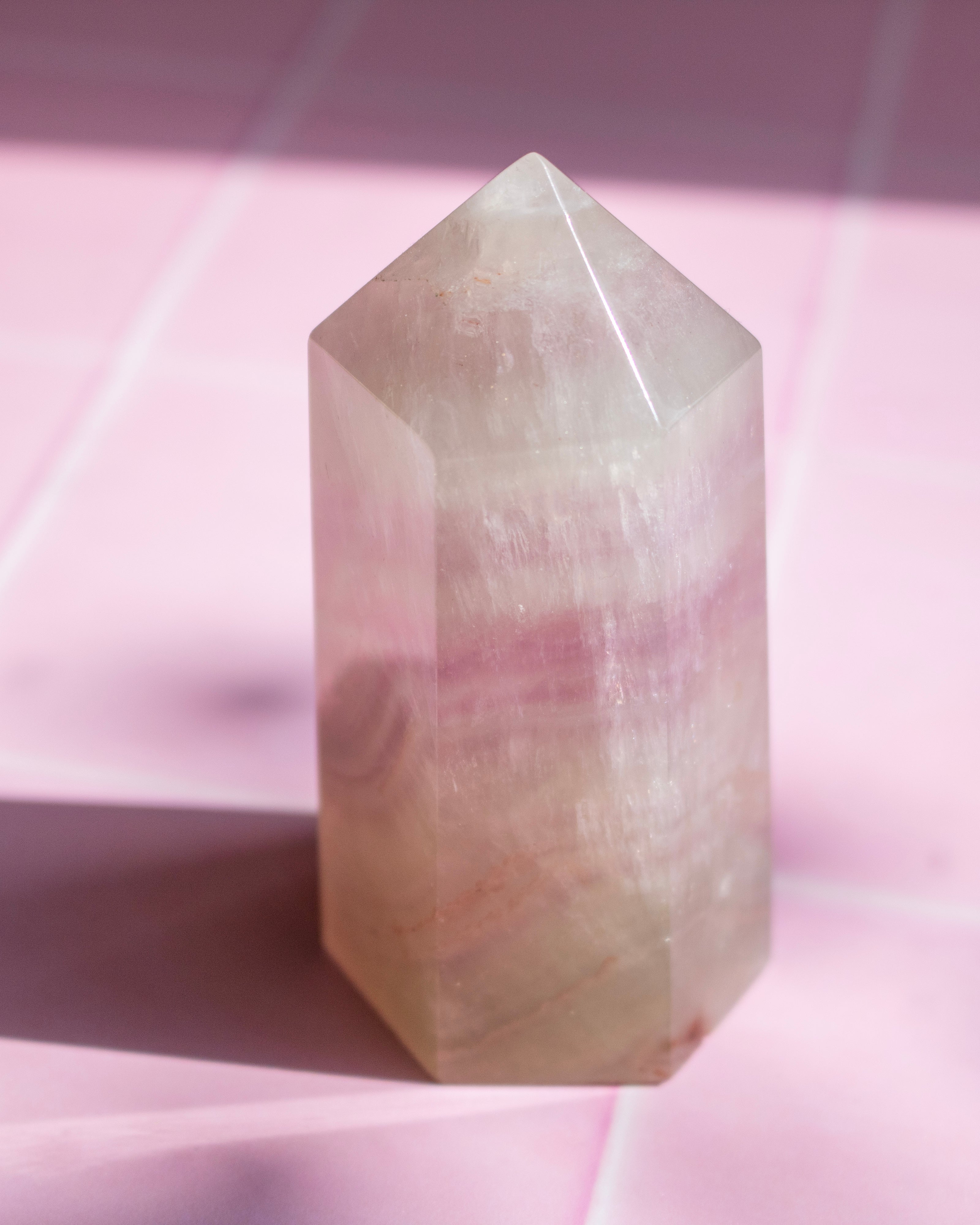 A polished natural crystal tower with pink and beige banding, standing on a pink tiled surface under direct sunlight.