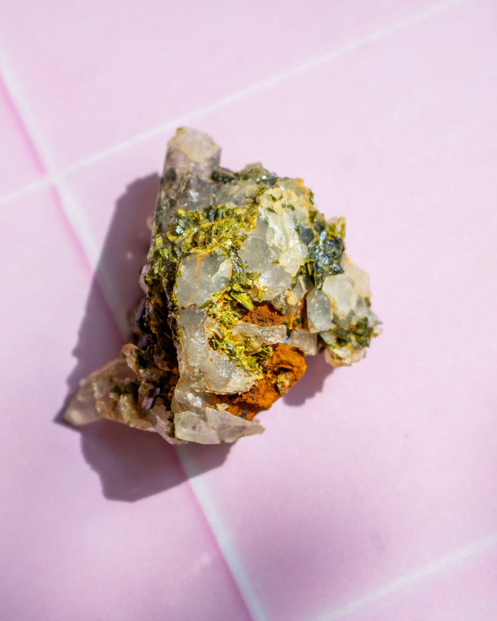 A raw mineral specimen with green, yellow, and orange crystalline textures on a light pink tiled background.