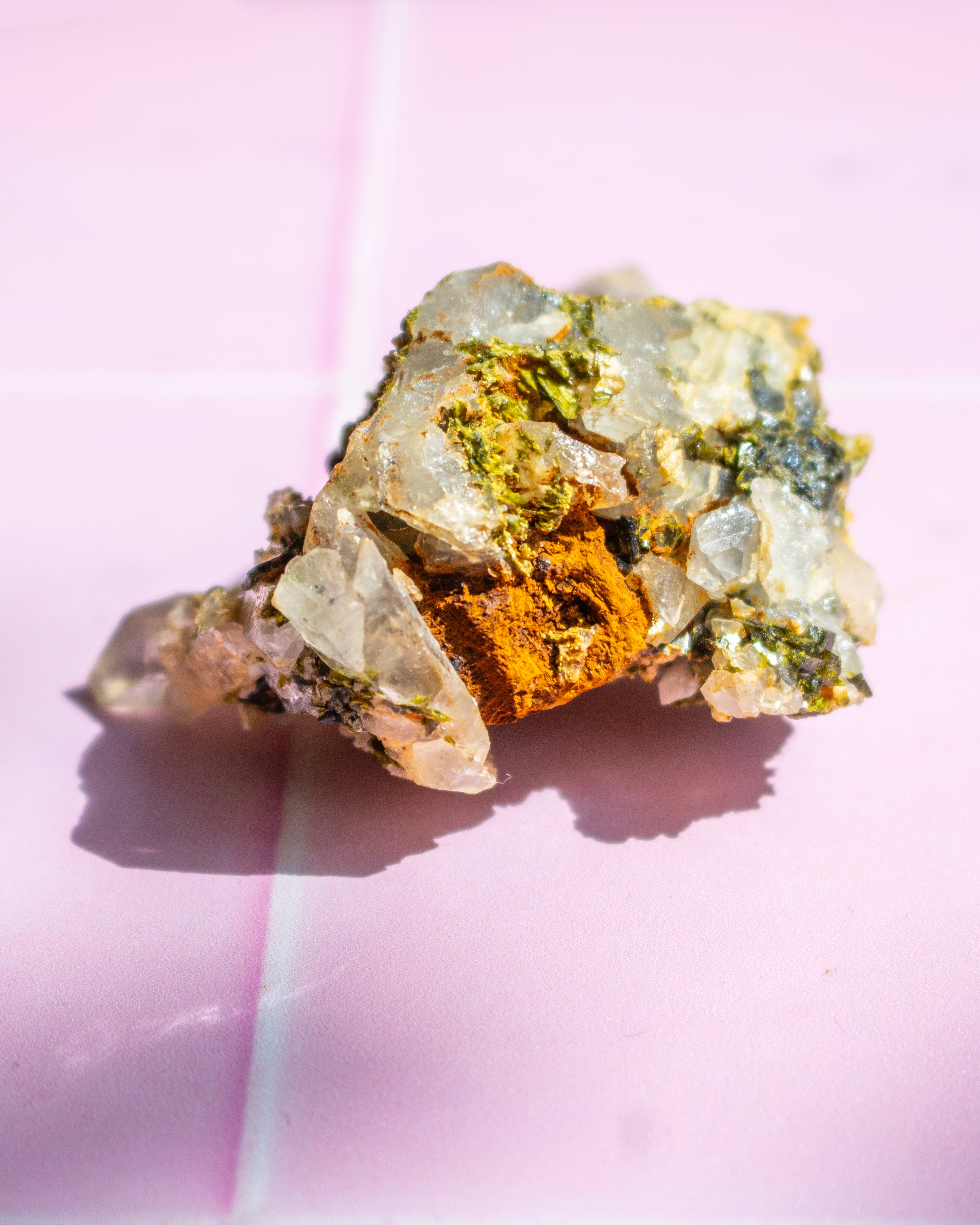 A raw mineral specimen with clear quartz crystals and a bright orange-brown metallic core, photographed on a pink background.