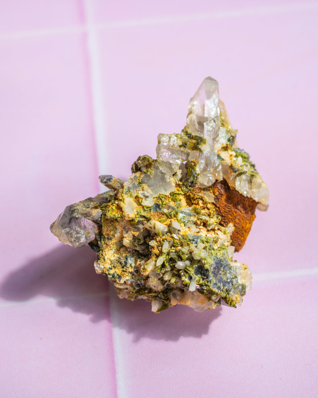 A natural mineral specimen featuring a cluster of clear quartz crystals over a matrix of green, yellow, and brown minerals, displayed on a pink tiled background.
