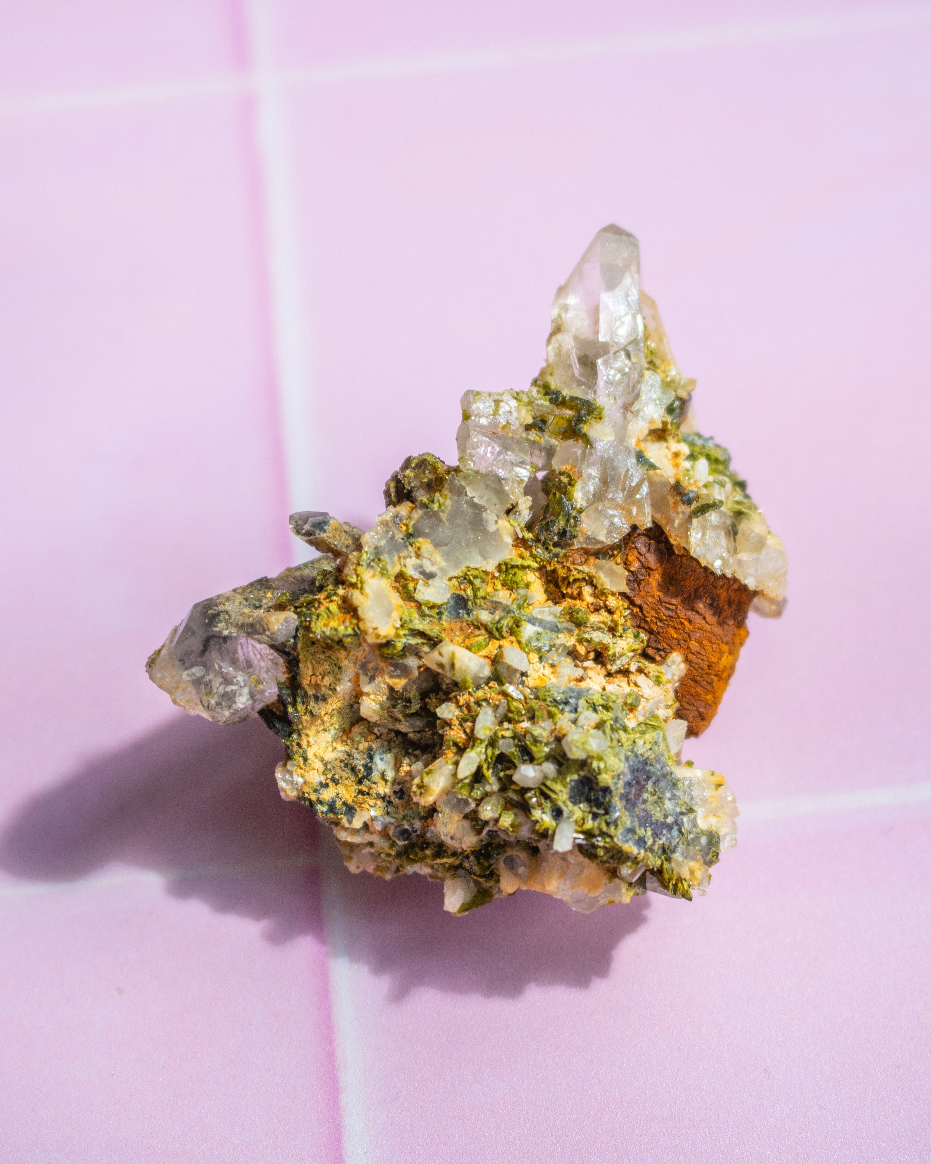 A natural mineral specimen featuring a cluster of clear quartz crystals over a matrix of green, yellow, and brown minerals, displayed on a pink tiled background.