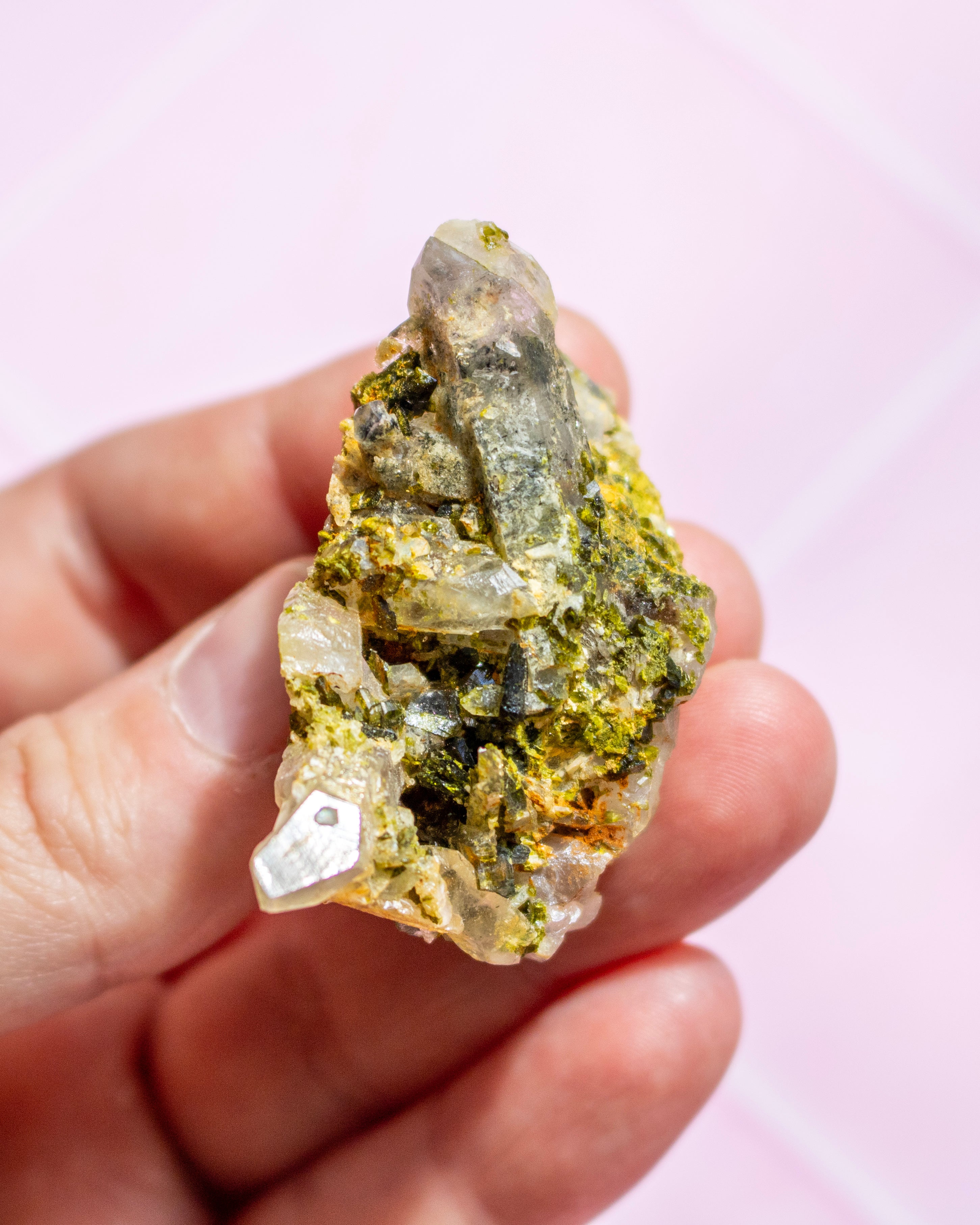 A hand holding a raw mineral specimen featuring a clear quartz crystal point over a yellow-green and dark gray matrix, set against a soft pink background.