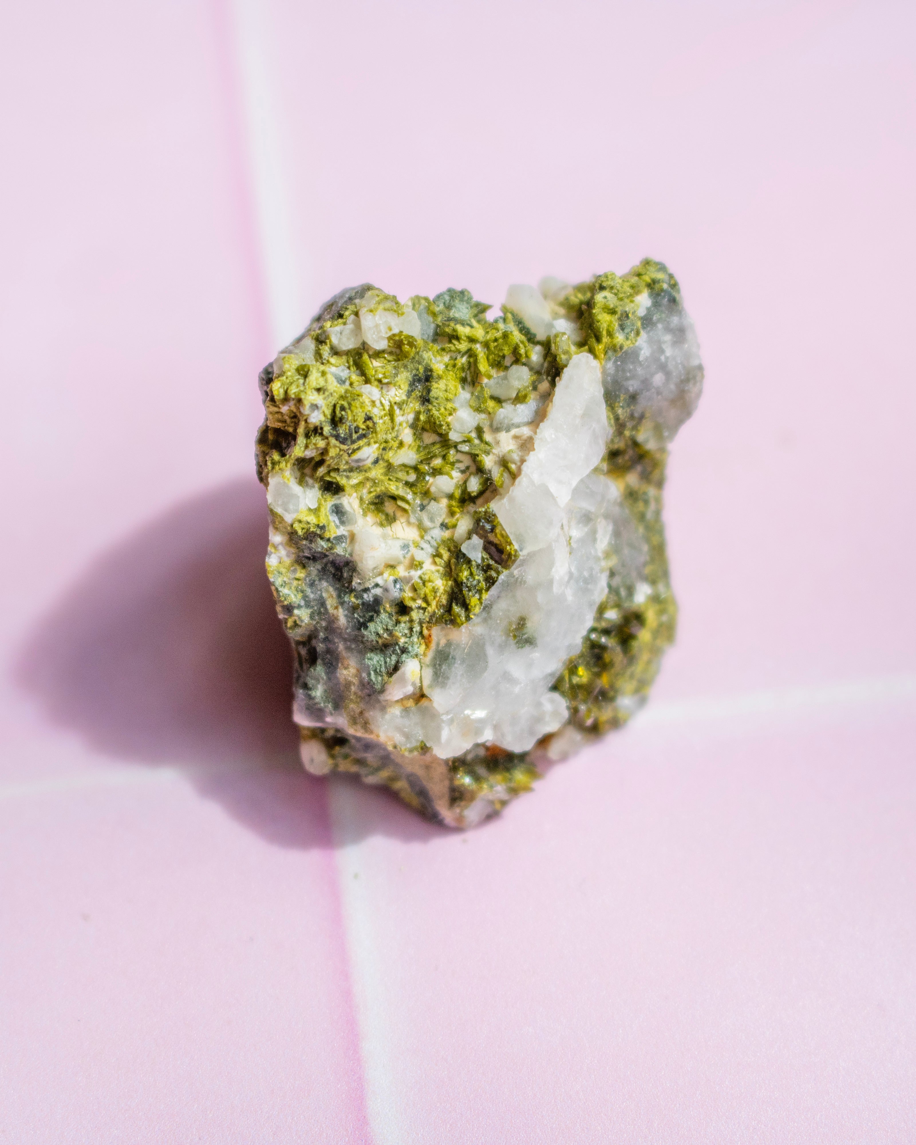A raw mineral specimen featuring a mix of green matrix, clear quartz crystals, and white quartz, photographed on a light pink background.