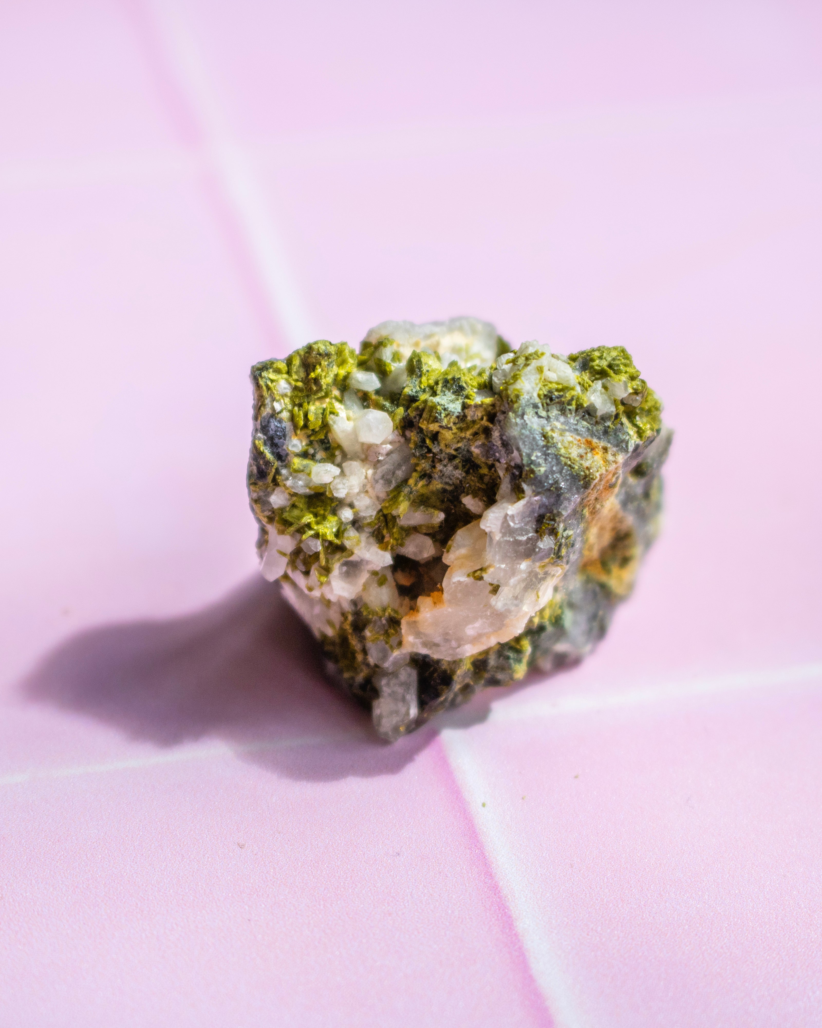 A small rough mineral or crystal specimen with green, gray, and translucent white crystalline areas, photographed on a pink surface with a visible shadow.