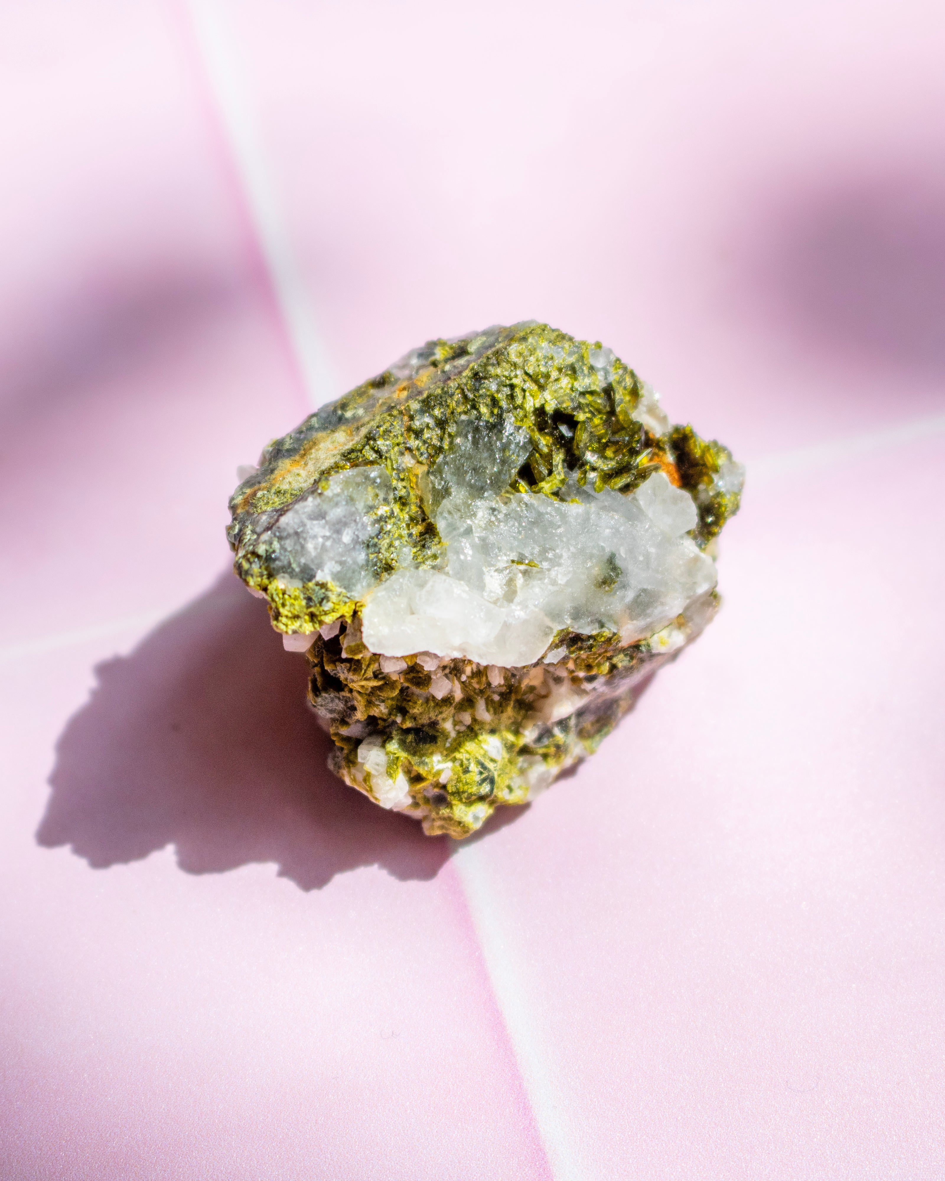 A raw mineral specimen with a bright green crystalline surface and a translucent white crystal section, photographed on a soft pink background with a cast shadow.