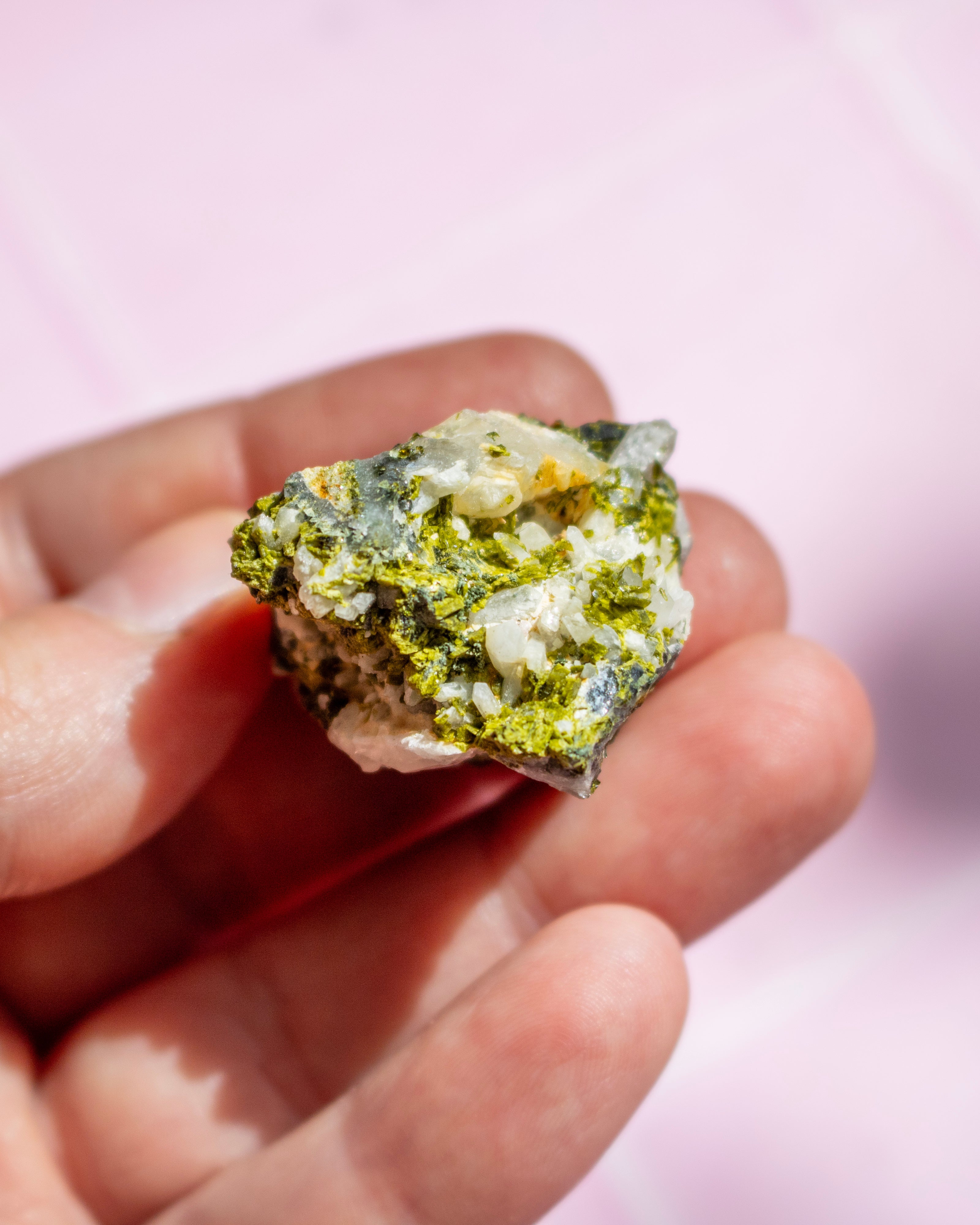 A hand holding a small, rough green and white mineral or crystal specimen against a soft pink background.