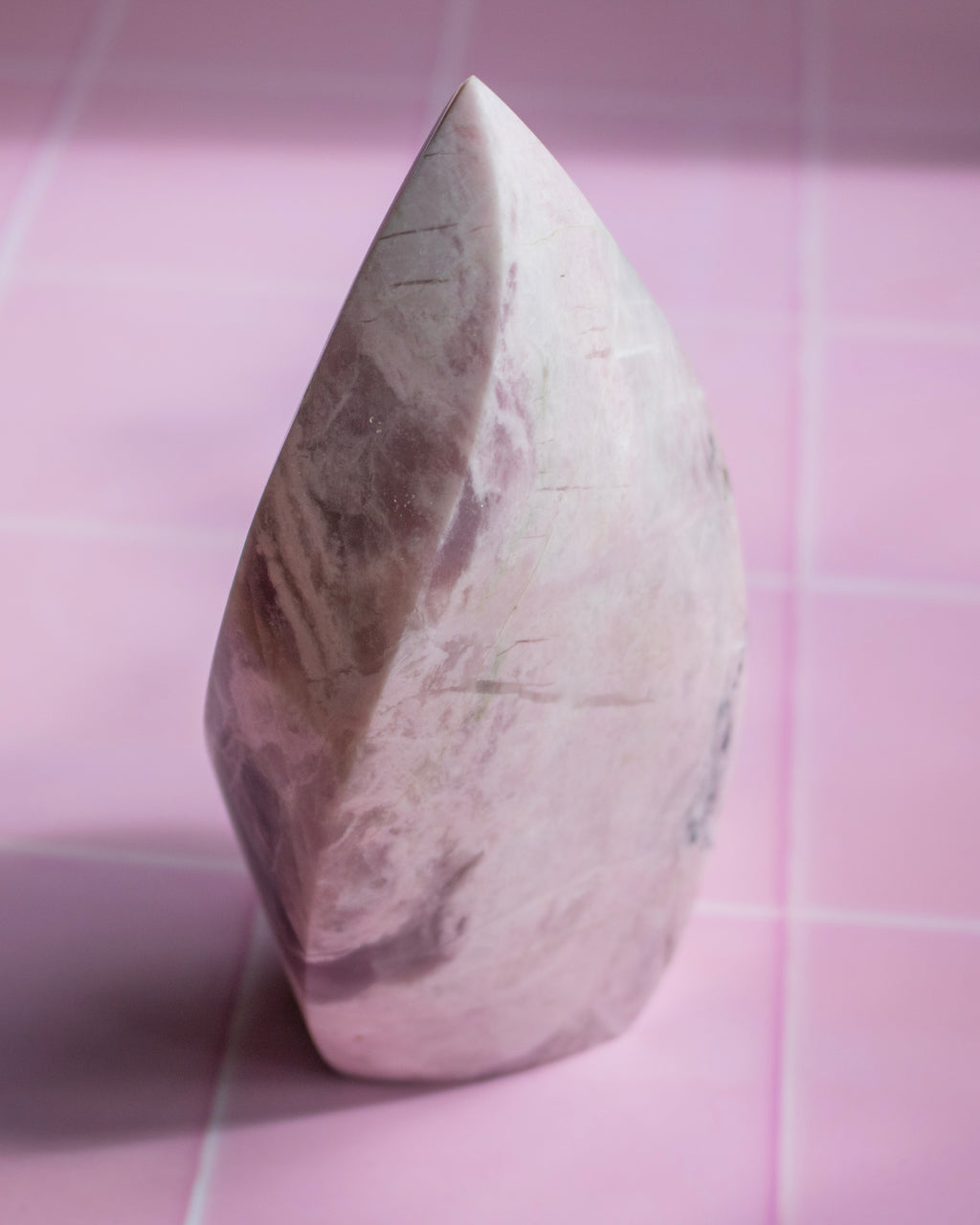 A polished teardrop-shaped crystal or stone ornament with natural pink, beige, and gray marbling, standing upright on a pink tiled surface.