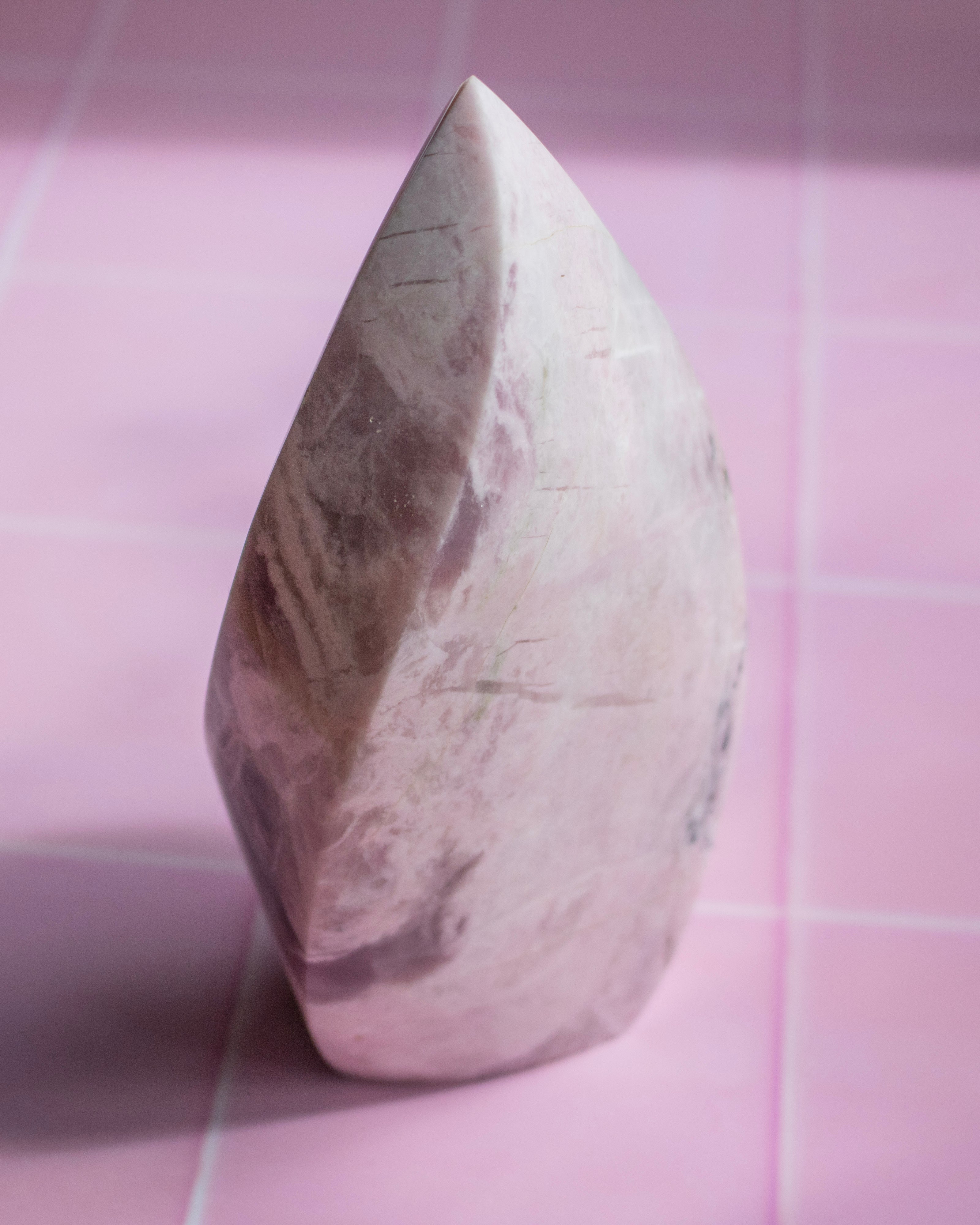 A polished teardrop-shaped crystal or stone ornament with natural pink, beige, and gray marbling, standing upright on a pink tiled surface.