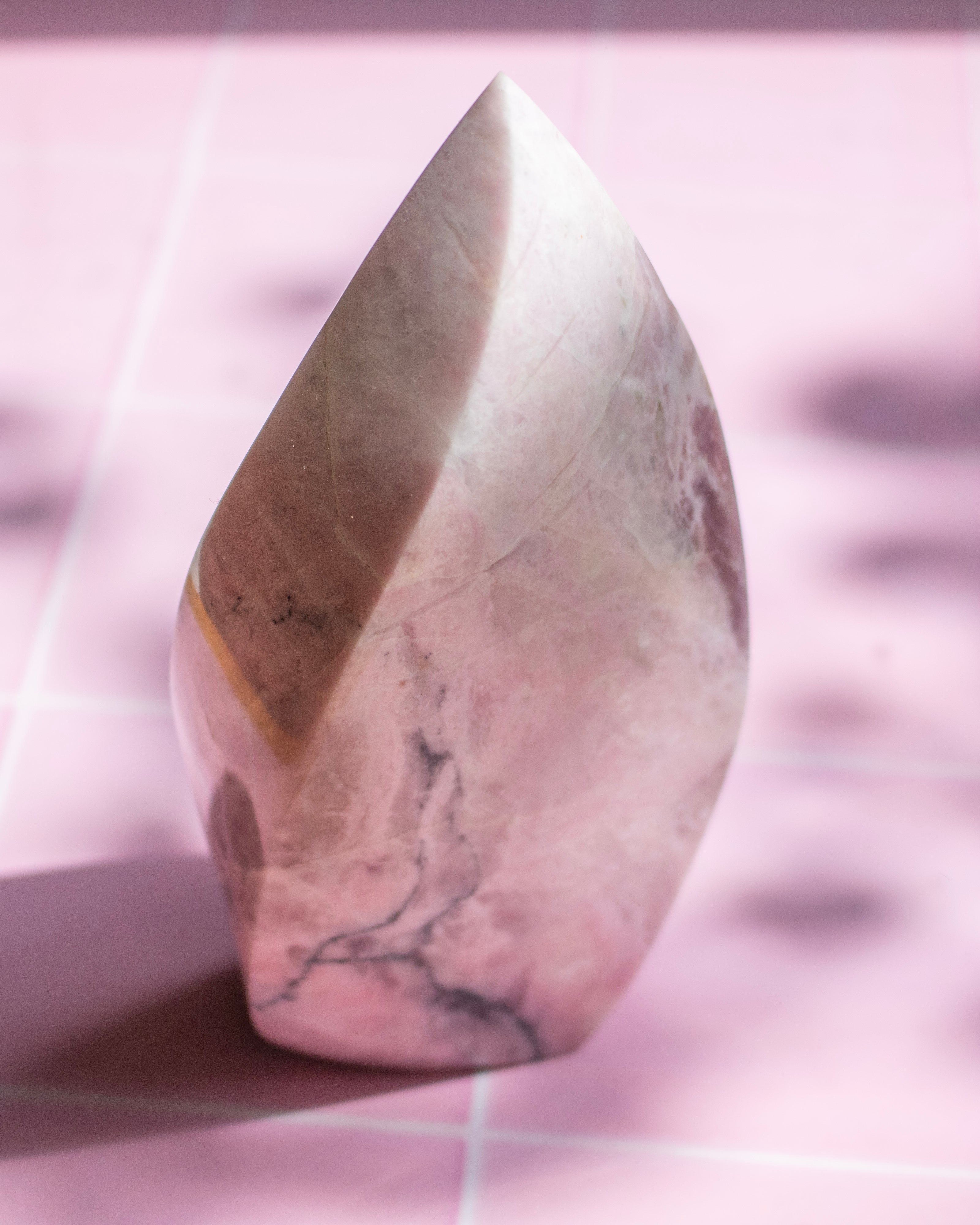 A polished pink stone crystal tower with natural marbled patterns, standing upright on a pink surface with a grid pattern background.