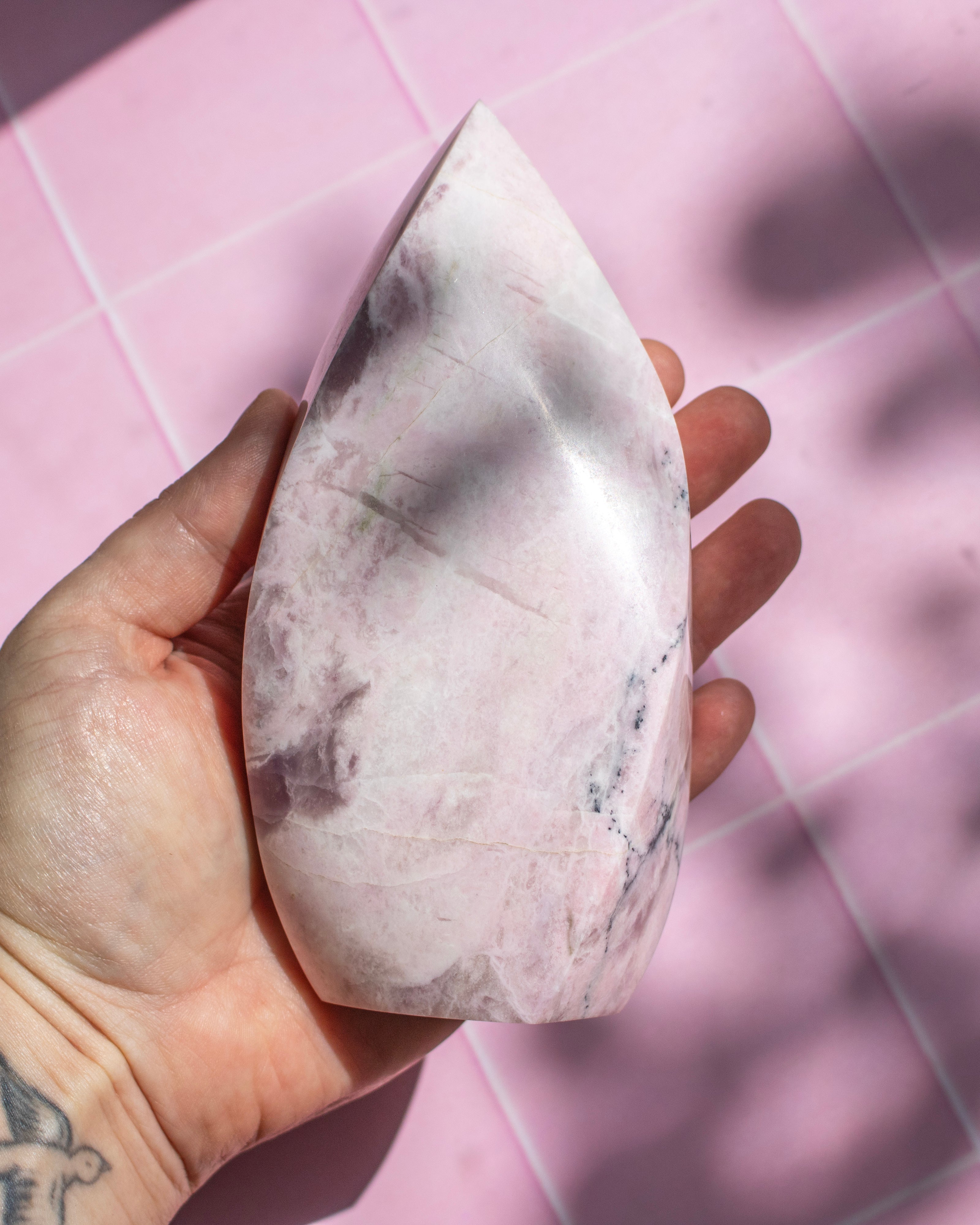 A hand holding a polished pink quartz crystal point against a pink tiled background.