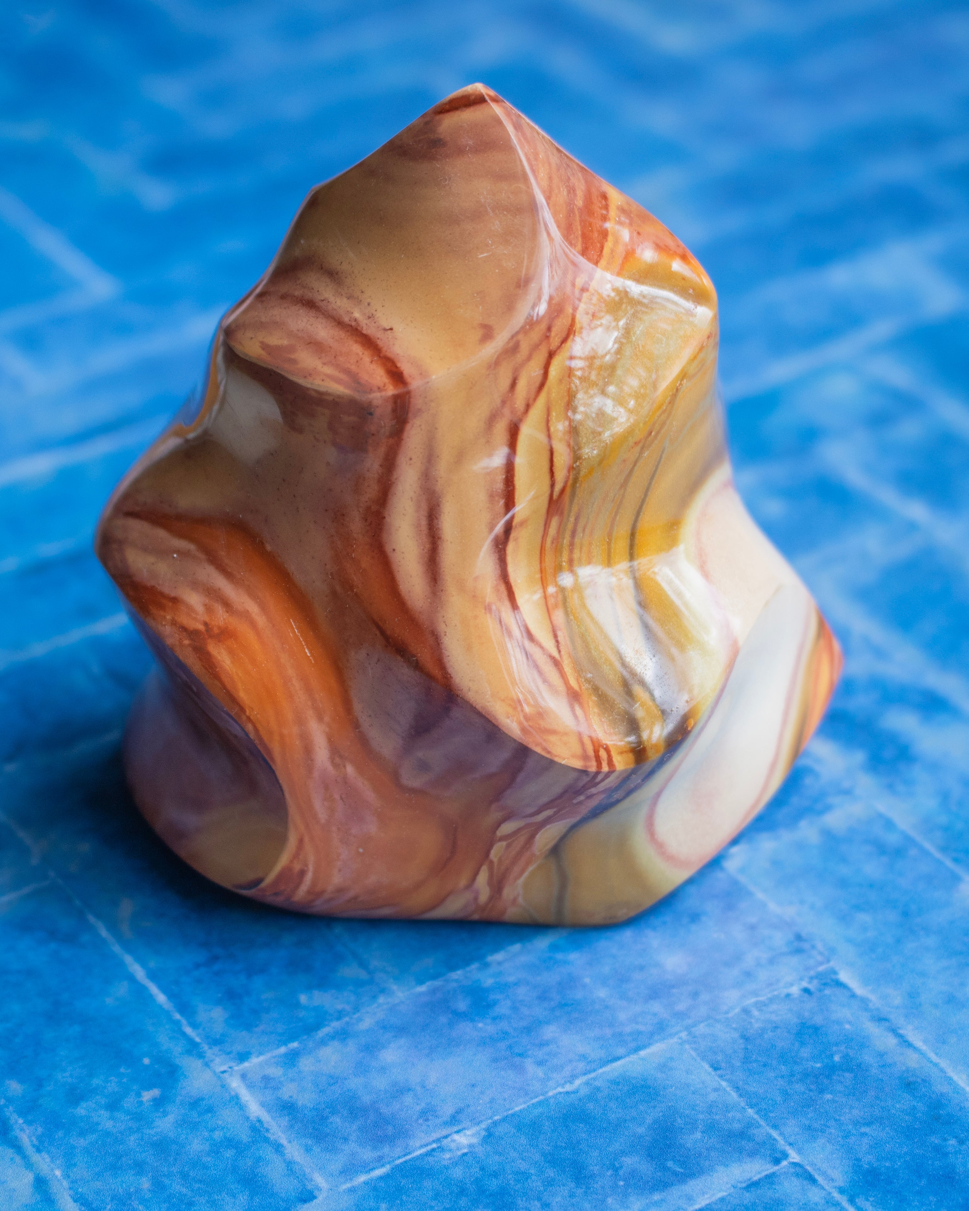 A polished decorative stone sculpture with swirling marbled patterns in orange, brown, beige, and cream, set against a textured blue background.