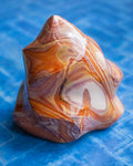 A polished crystal with swirling bands of orange, brown, cream, and purple, displayed on a textured blue surface.