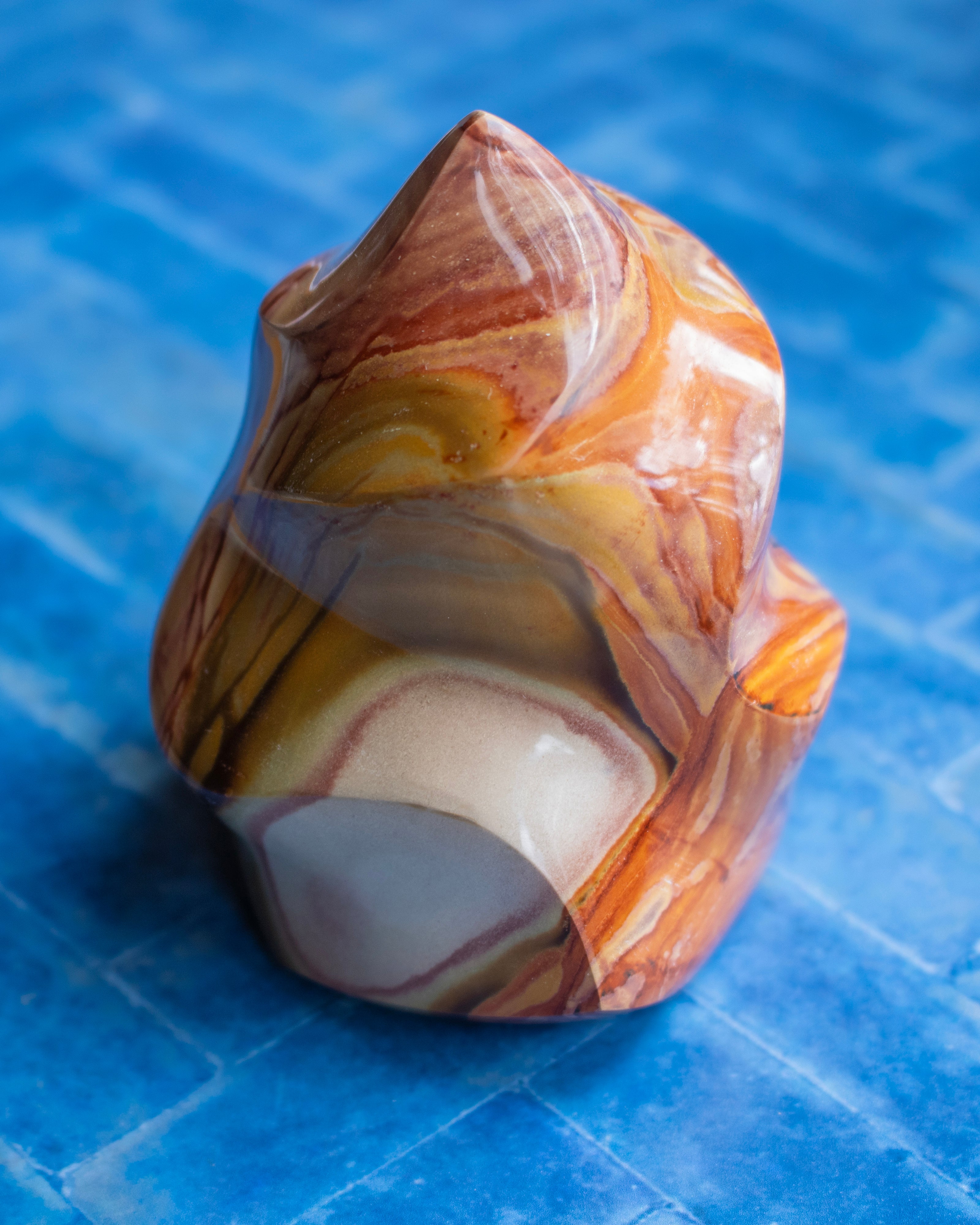 A polished carved stone sculpture with swirling marbled patterns in orange, brown, cream, and yellow, set against a bright blue tiled background.
