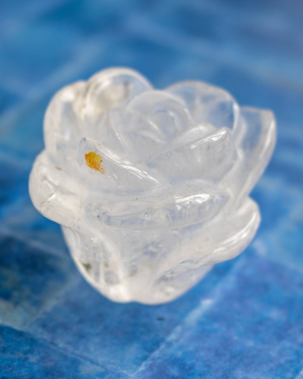 A small carved translucent white rose-shaped object with a tiny yellow inclusions or spot, photographed close-up against a blue textured background.