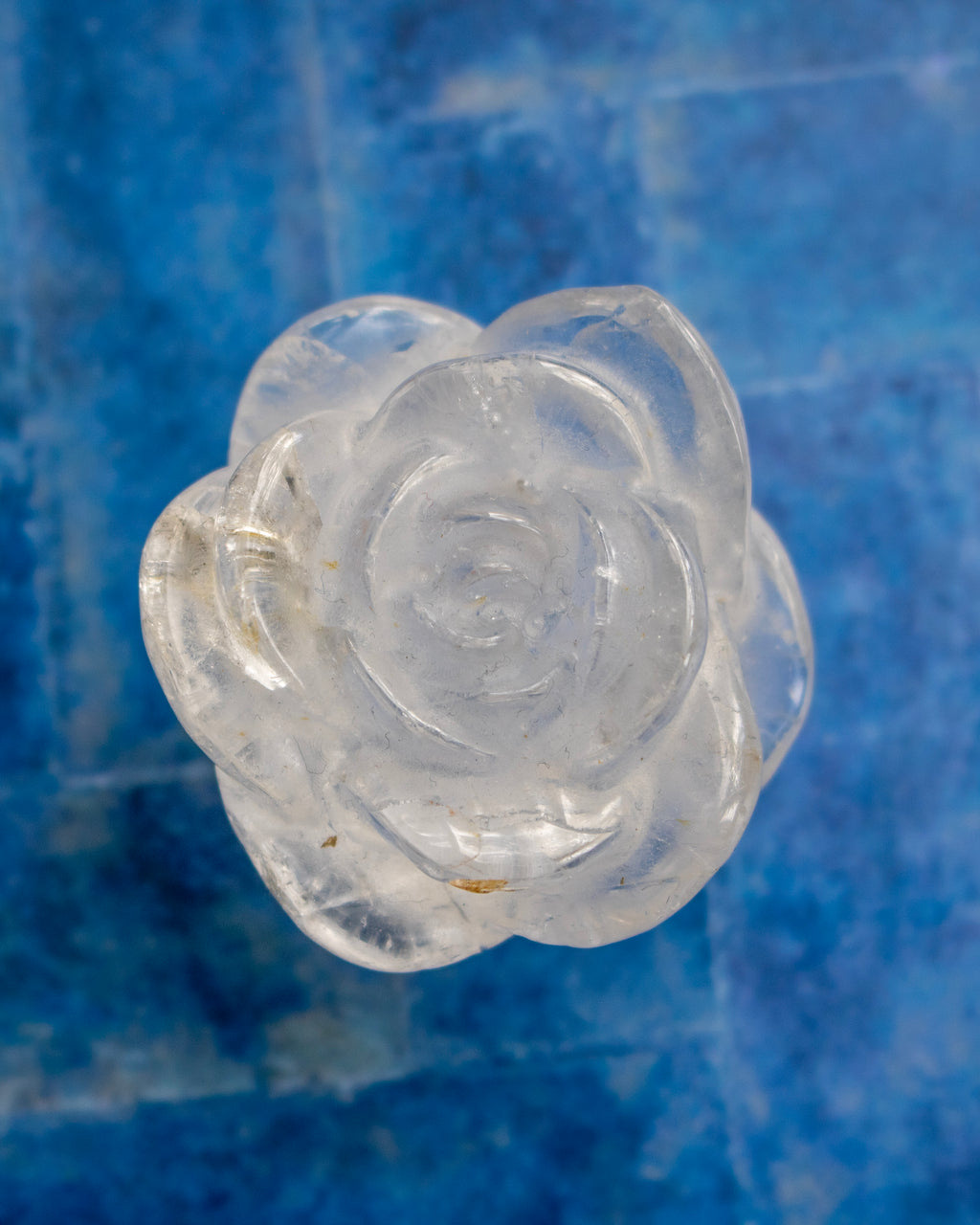 A clear, translucent carved rose-shaped object, possibly a crystal or glass decorative piece, shown against a textured blue background.