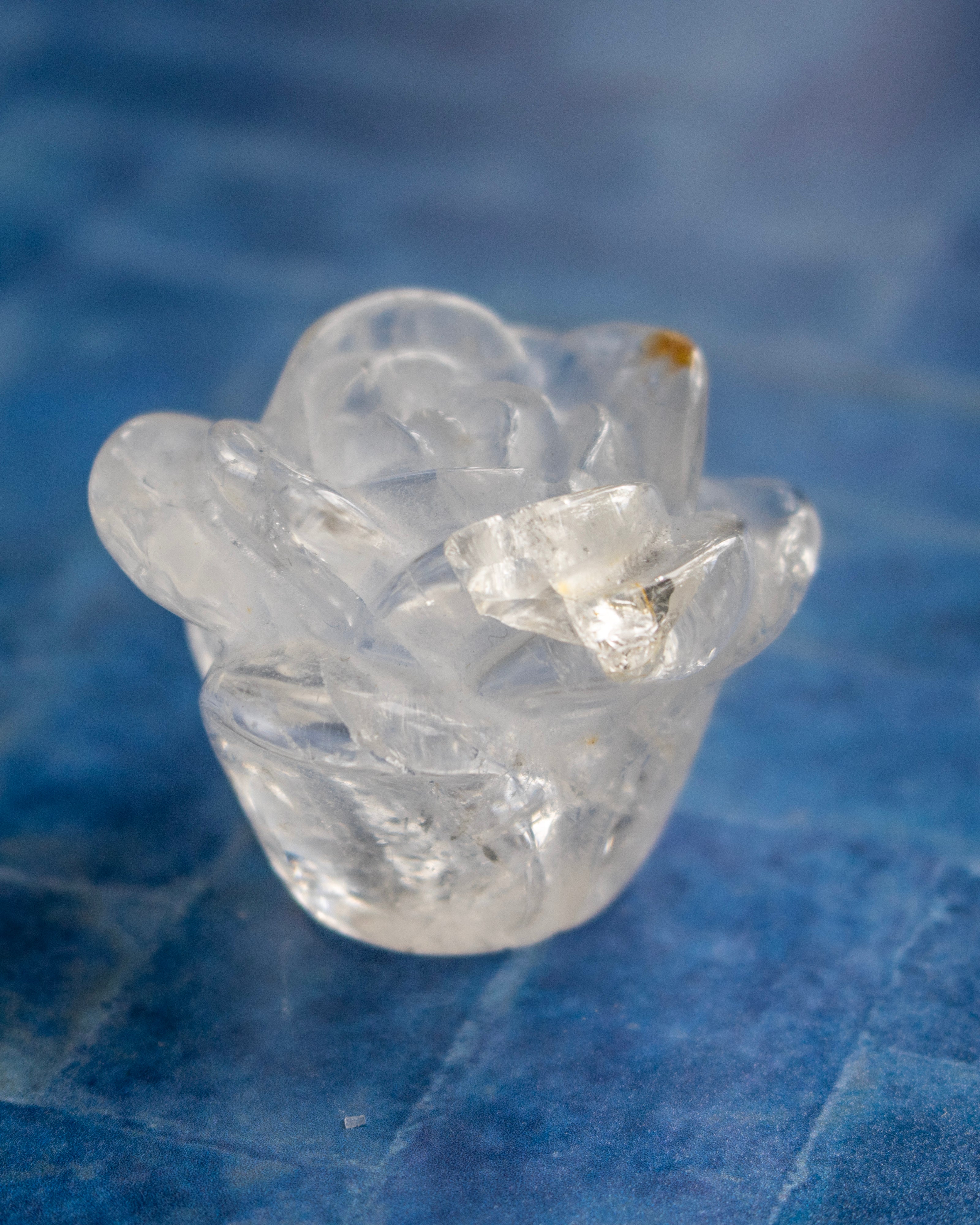 A small translucent clear crystal rose figurine with a textured, layered petal design, resting on a blue textured surface.